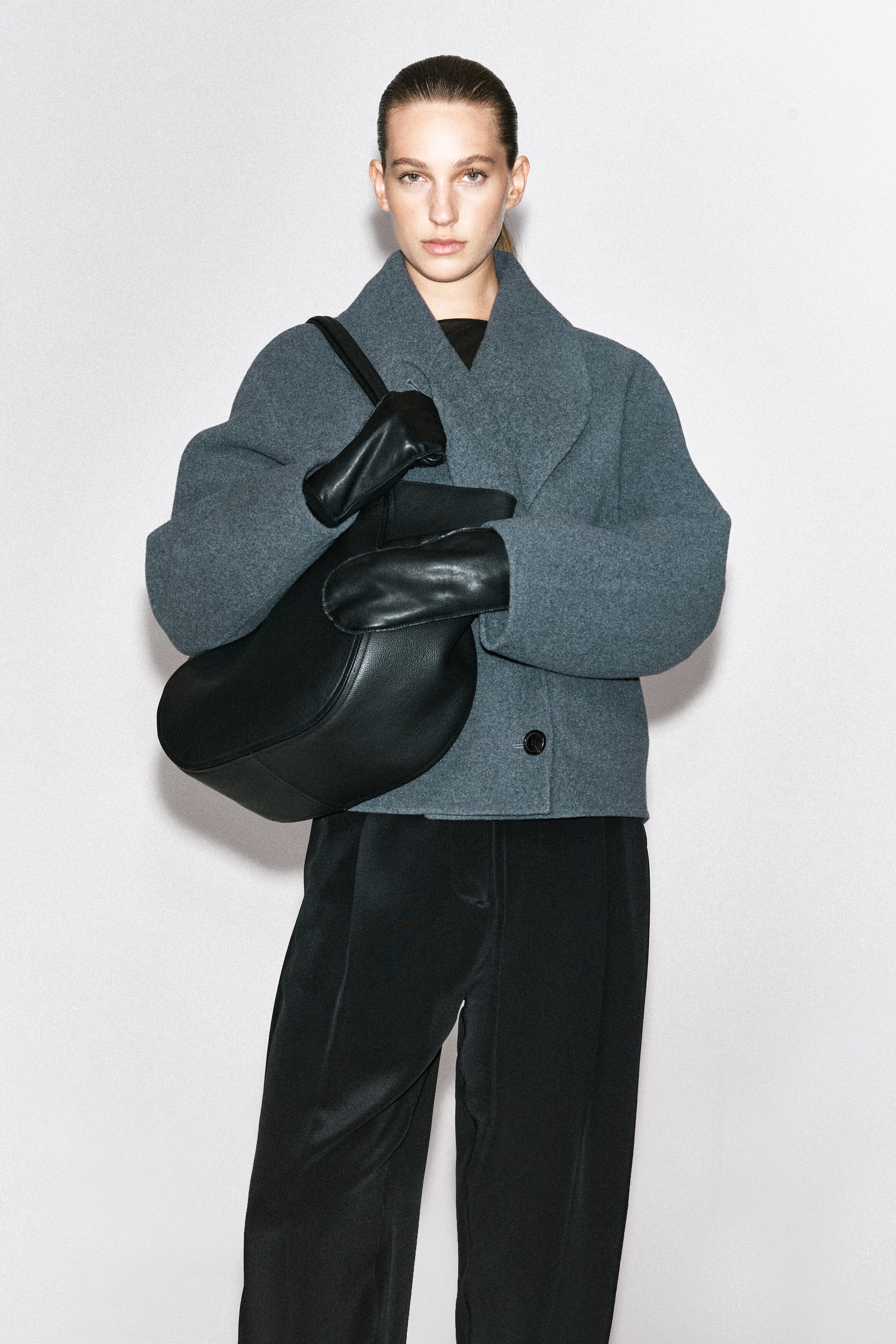 A cropped gray wool jacket with a wide shawl collar and a single-button closure is worn by the model. A large black leather shoulder bag is carried and held with black leather gloves, styled with black trousers. A cropped gray wool jacket with a wide shawl collar and a single-button closure is worn by the model. A large black leather shoulder bag is carried and held with black leather gloves, styled with black trousers.