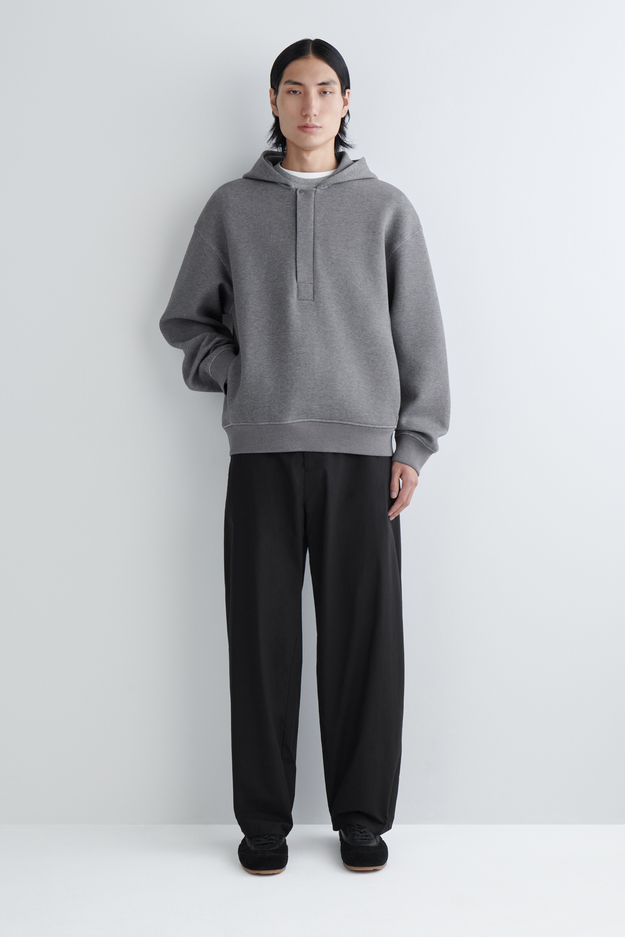 Zobrazit větší obrázek: Šedá mikina s kapucí je prezentována zepředu, má volný střih, krátkou falešnou légu pod výstřihem a viditelnou kapuci. Má žebrované manžety a žebrovaný lem a je stylizována přes bílé tričko, v kombinaci s černými širokými kalhotami a černými nízkými teniskami.