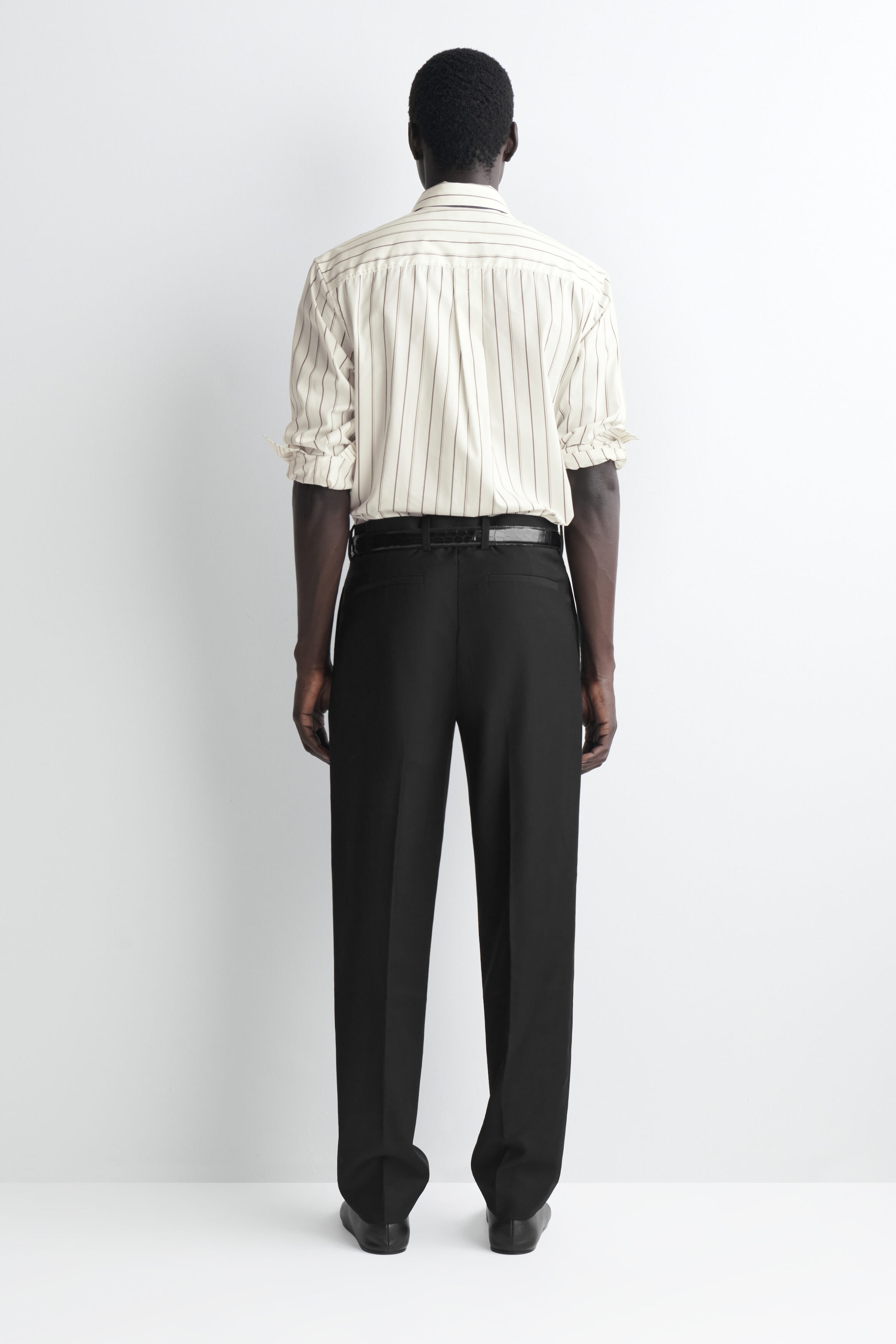 Black straight-leg trousers are shown from the back, featuring a regular fit, pressed creases down each leg, and two rear welt pockets. The trousers are styled with a tucked-in striped shirt and a black belt, emphasising their clean waistband structure. Black straight-leg trousers are shown from the back, featuring a regular fit, pressed creases down each leg, and two rear welt pockets. The trousers are styled with a tucked-in striped shirt and a black belt, emphasising their clean waistband structure.