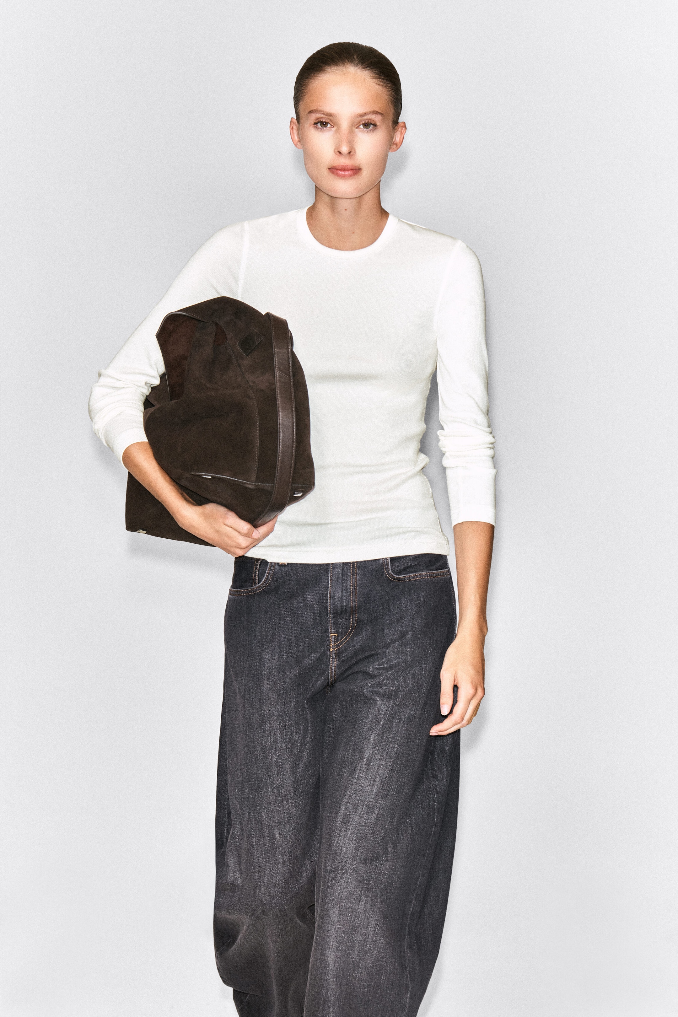 A white, slim-ribbed cotton long-sleeve top with a crew neck is worn by the model. The top is styled with dark gray wide-leg jeans, and a large dark brown suede bag is carried under one arm. A white, slim-ribbed cotton long-sleeve top with a crew neck is worn by the model. The top is styled with dark gray wide-leg jeans, and a large dark brown suede bag is carried under one arm.