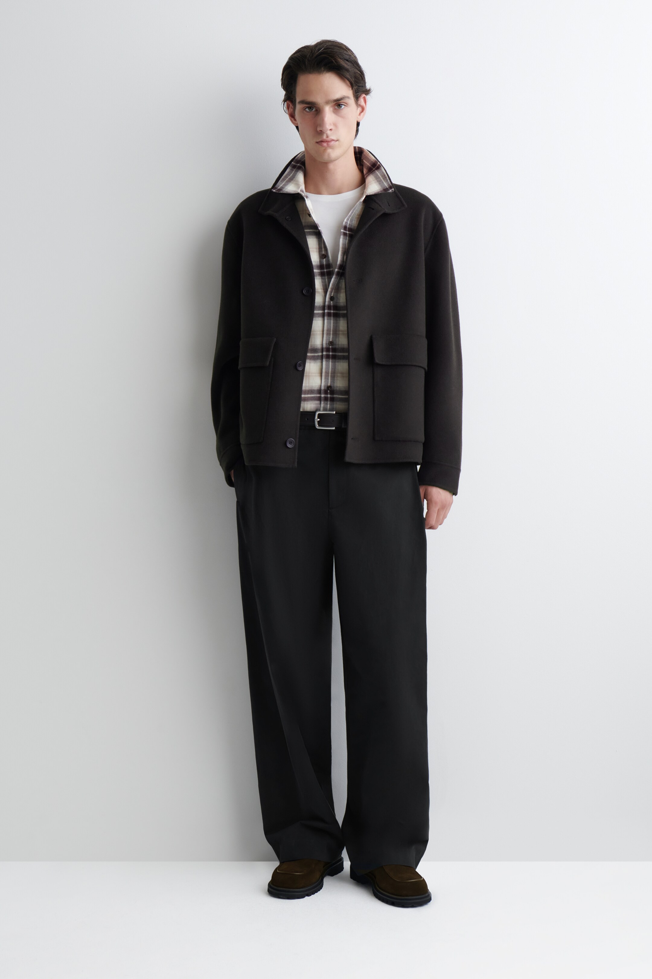 View larger image: A front-facing outfit is being presented, featuring a dark, short jacket with a collar and large flap pockets, which is being worn open. A plaid shirt is being layered underneath, over a white crew-neck t-shirt. Dark, wide-leg trousers are being worn, cinched with a dark belt, and dark, thick-soled shoes are completing the look.