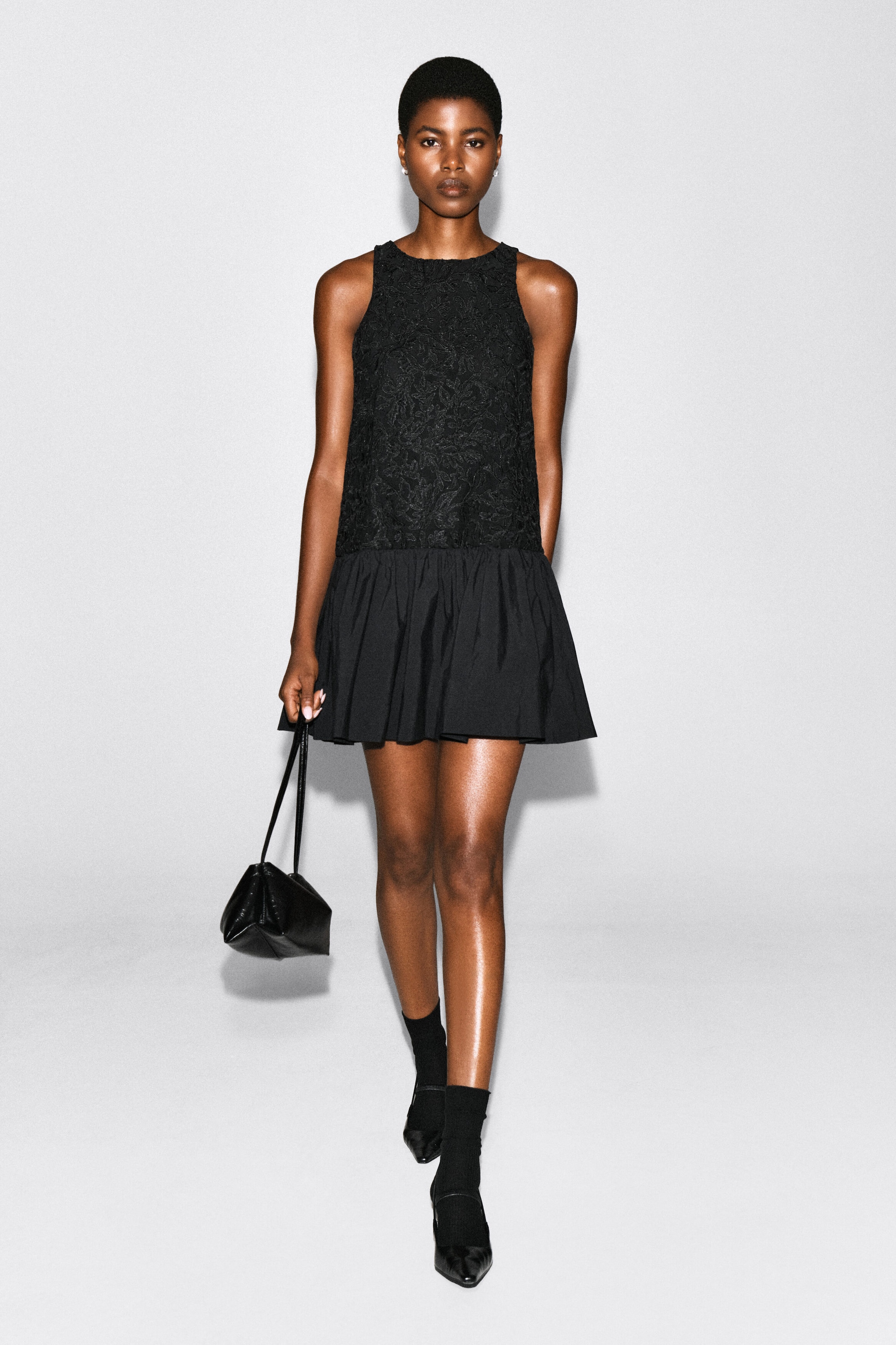 A black sleeveless mini dress featuring a textured, floral-embroidered bodice and a gathered, voluminous poplin skirt is being worn by the model. The model is walking forwards while carrying a small black handbag and wearing matching black socks with pointed-toe shoes. A black sleeveless mini dress featuring a textured, floral-embroidered bodice and a gathered, voluminous poplin skirt is being worn by the model. The model is walking forwards while carrying a small black handbag and wearing matching black socks with pointed-toe shoes.