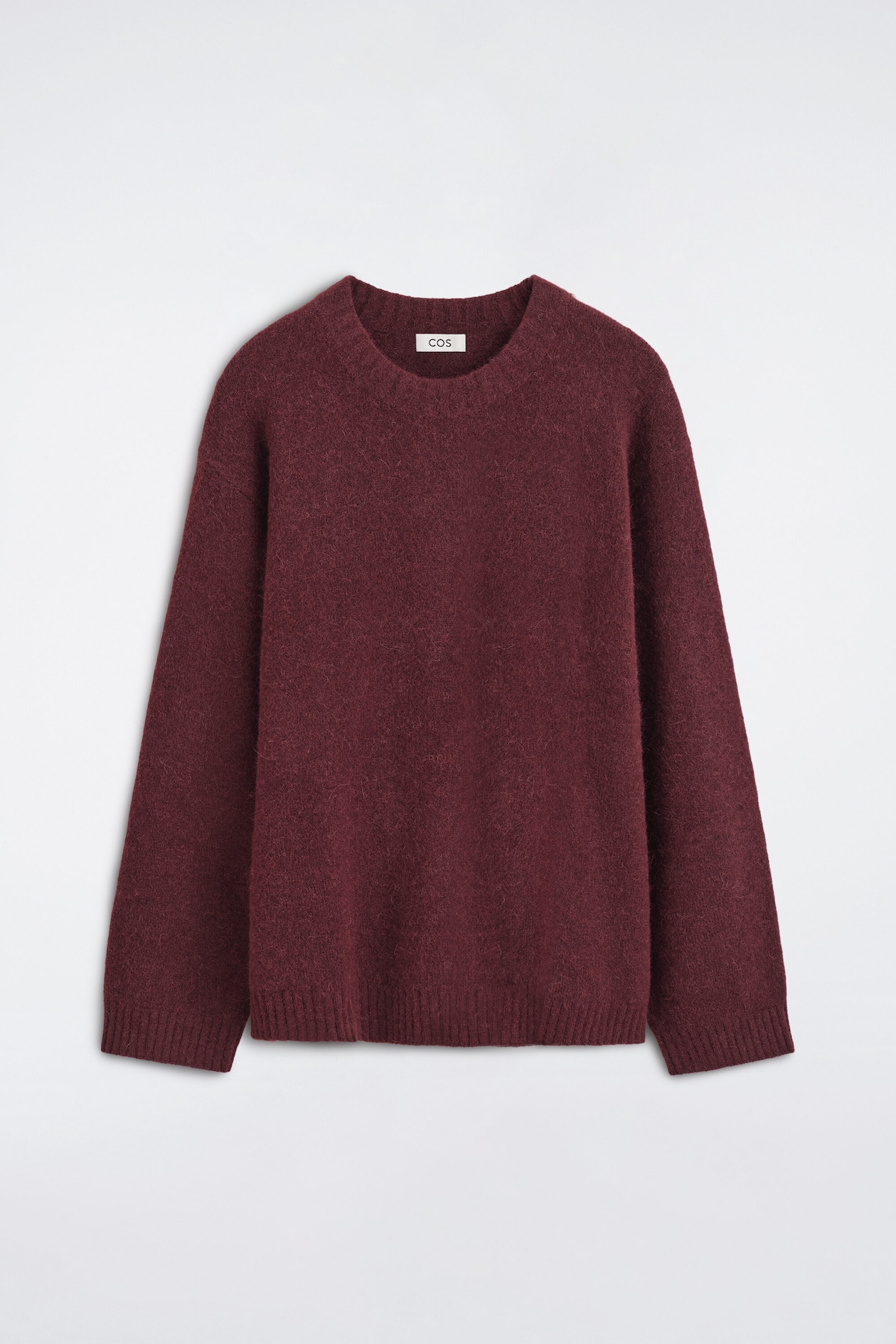 An oversized alpaca-blend crew-neck sweater in a deep burgundy hue presents a relaxed, loose-fitting silhouette from the front. The garment features a fuzzy, textured knit with a ribbed crew neck, dropped shoulders, ribbed cuffs, and a ribbed hem.