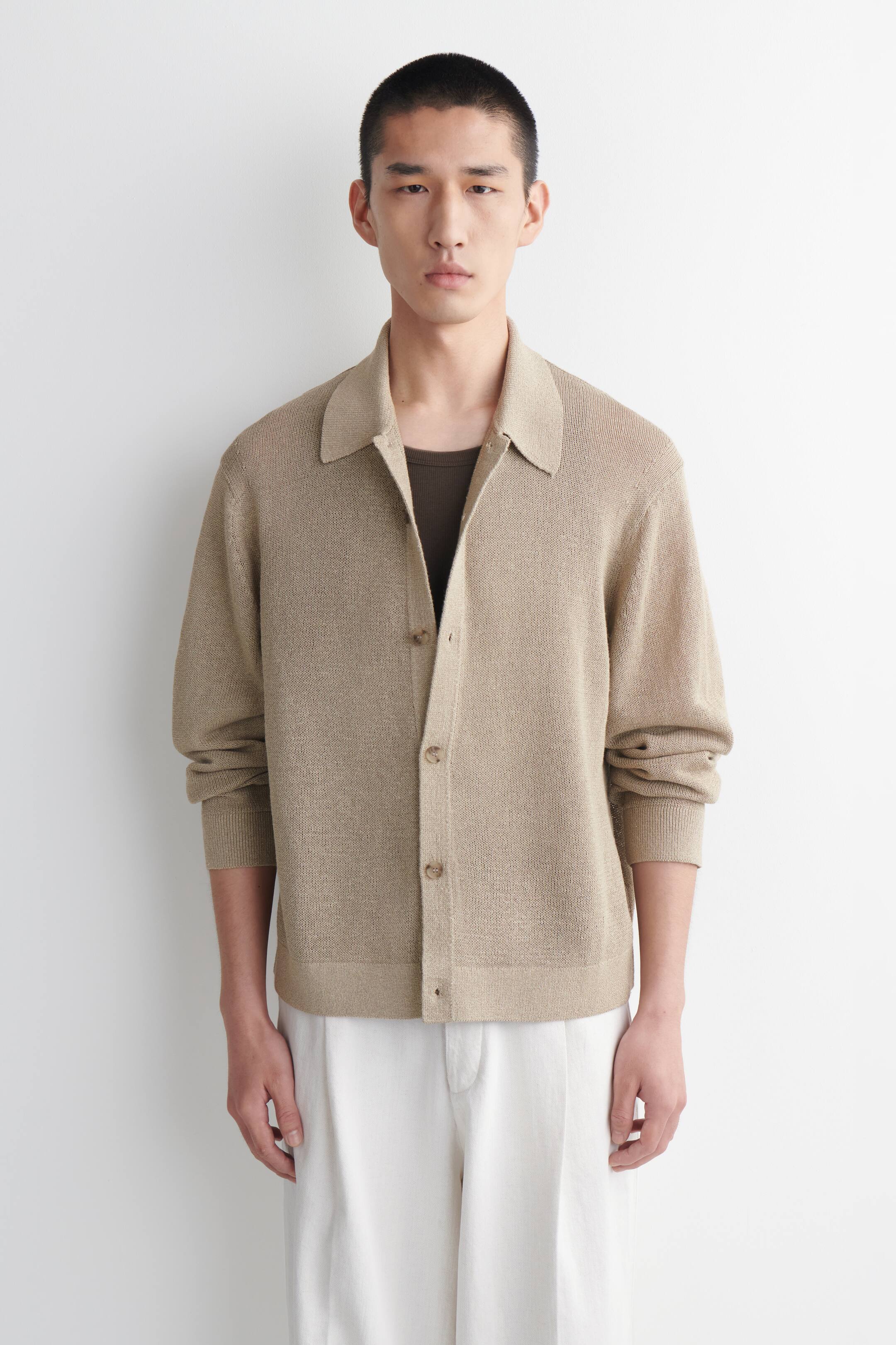 Agrandir l'image: Une chemise beige en tricot à boutons est dotée d'un col classique à pointes, de manches longues et d'un ourlet droit. Son devant est partiellement boutonné avec des boutons marron clair, portée sur un t-shirt foncé et associée à un pantalon plissé blanc.