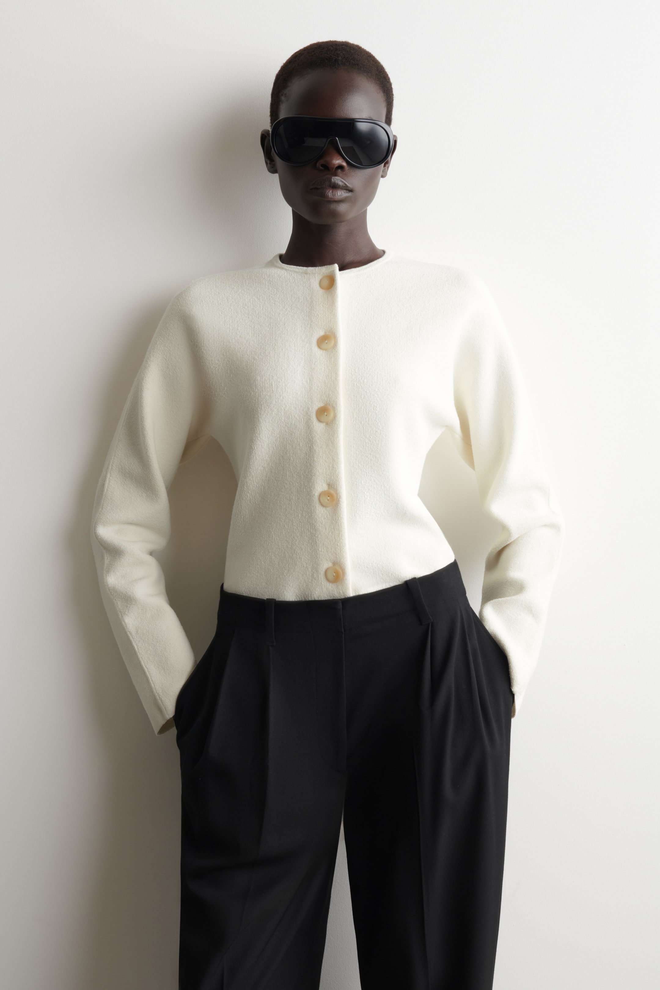הצגת תמונה גדולה יותר: קרדיגן בצבע קרם מוצג מקדימה, עם מחשוף עגול וחמישה כפתורים חומים בהירים הסוגרים את החלק הקדמי. הוא מציג שרוולים ארוכים וגזרה רפויה וקצרה, המסתיימת במותן ומעוצבת מעל מכנסיים שחורים.