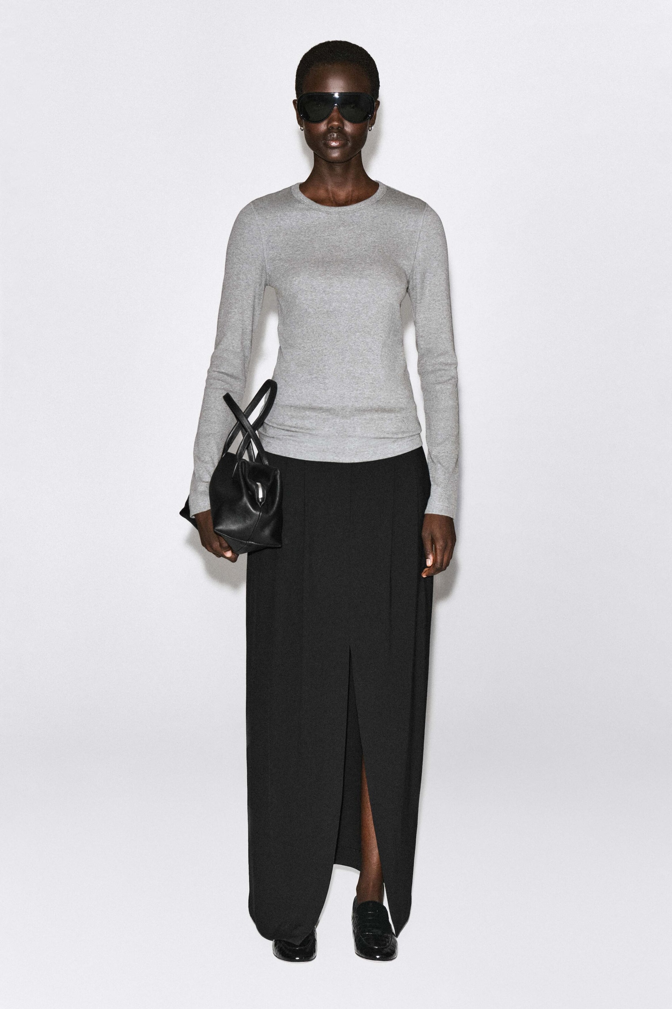 A slim ribbed cotton long-sleeved top in light grey is shown from the front, featuring a crew neckline and a close-fitting silhouette. The top extends to the hips and is styled with a black maxi skirt to highlight its slim fit. A slim ribbed cotton long-sleeved top in light grey is shown from the front, featuring a crew neckline and a close-fitting silhouette. The top extends to the hips and is styled with a black maxi skirt to highlight its slim fit.