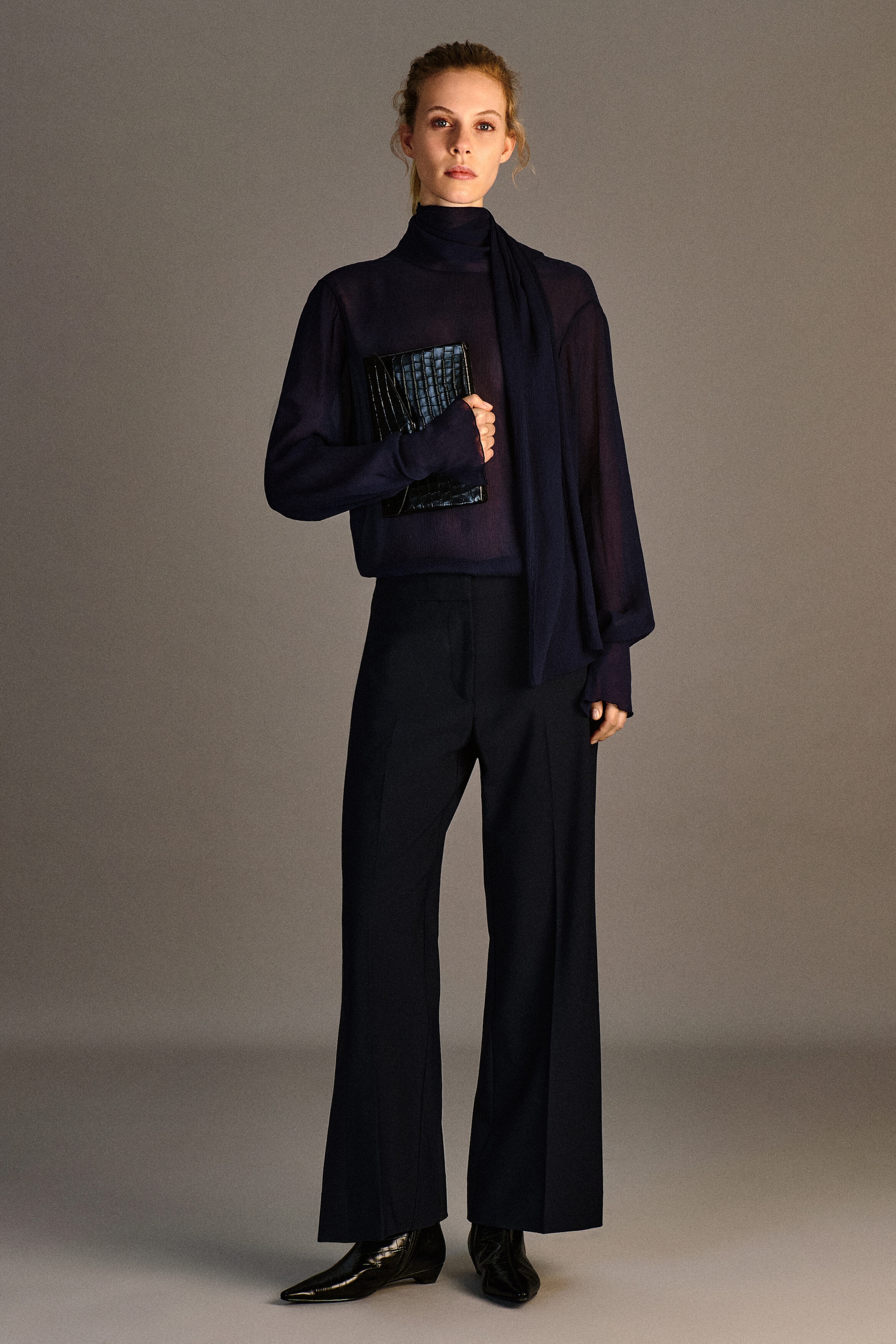 A model is standing in a front view, wearing a dark navy scarf-detail sheer crepe blouse tucked into matching twill cropped flared pants. The sheer, long-sleeved blouse features a high collar with an attached long scarf draped over the shoulder, while the high-waisted trousers showcase a structured wide-leg fit with sharp front creases.