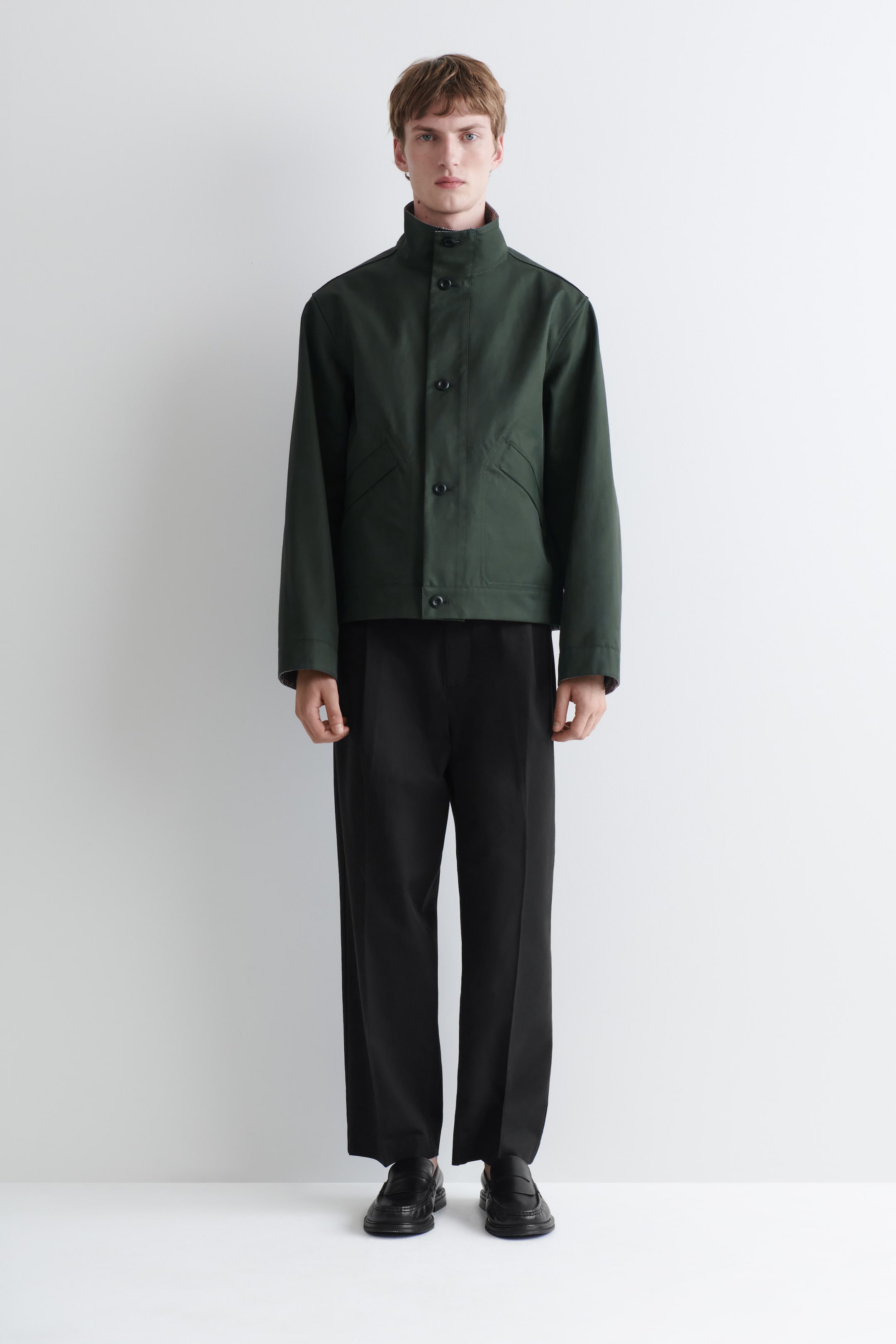 Größeres Bild anzeigen: Eine dunkelgrüne Jacke mit Stehkragen, Knopfleiste vorne und zwei schrägen Fronttaschen. Sie wird nach vorne getragen und mit einer schwarzen Wide-Leg-Hose kombiniert, die eine lockere Passform, Bundfalten vorne und eine verkürzte Länge aufweist.