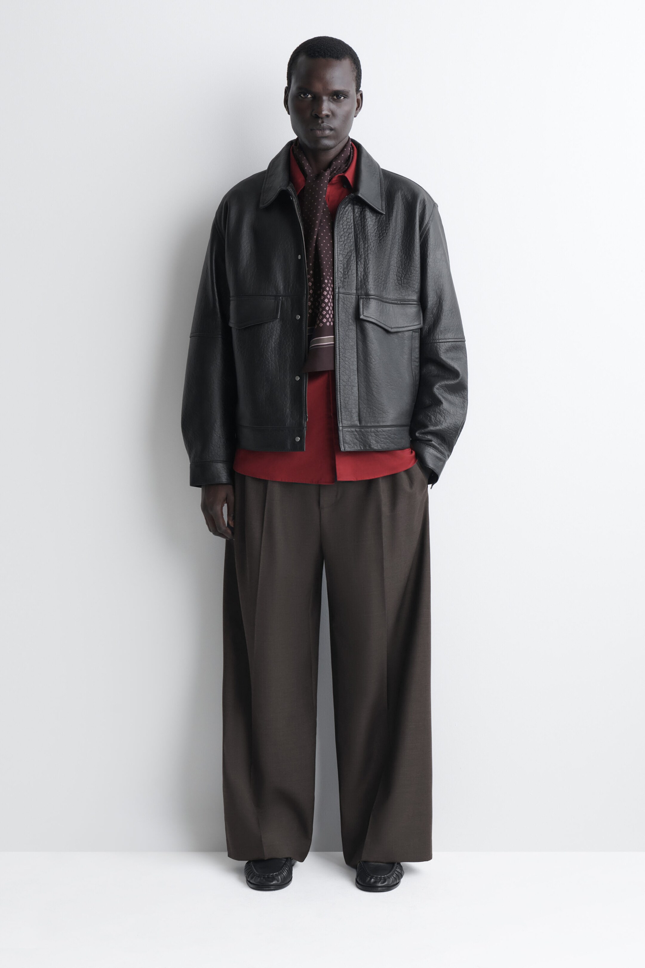 A slim, dark brown silk scarf featuring a micro diamond motif is shown from the front, draped around the neck and hanging down the chest. The scarf is layered under a red collared shirt and an open black leather jacket. A slim, dark brown silk scarf featuring a micro diamond motif is shown from the front, draped around the neck and hanging down the chest. The scarf is layered under a red collared shirt and an open black leather jacket.