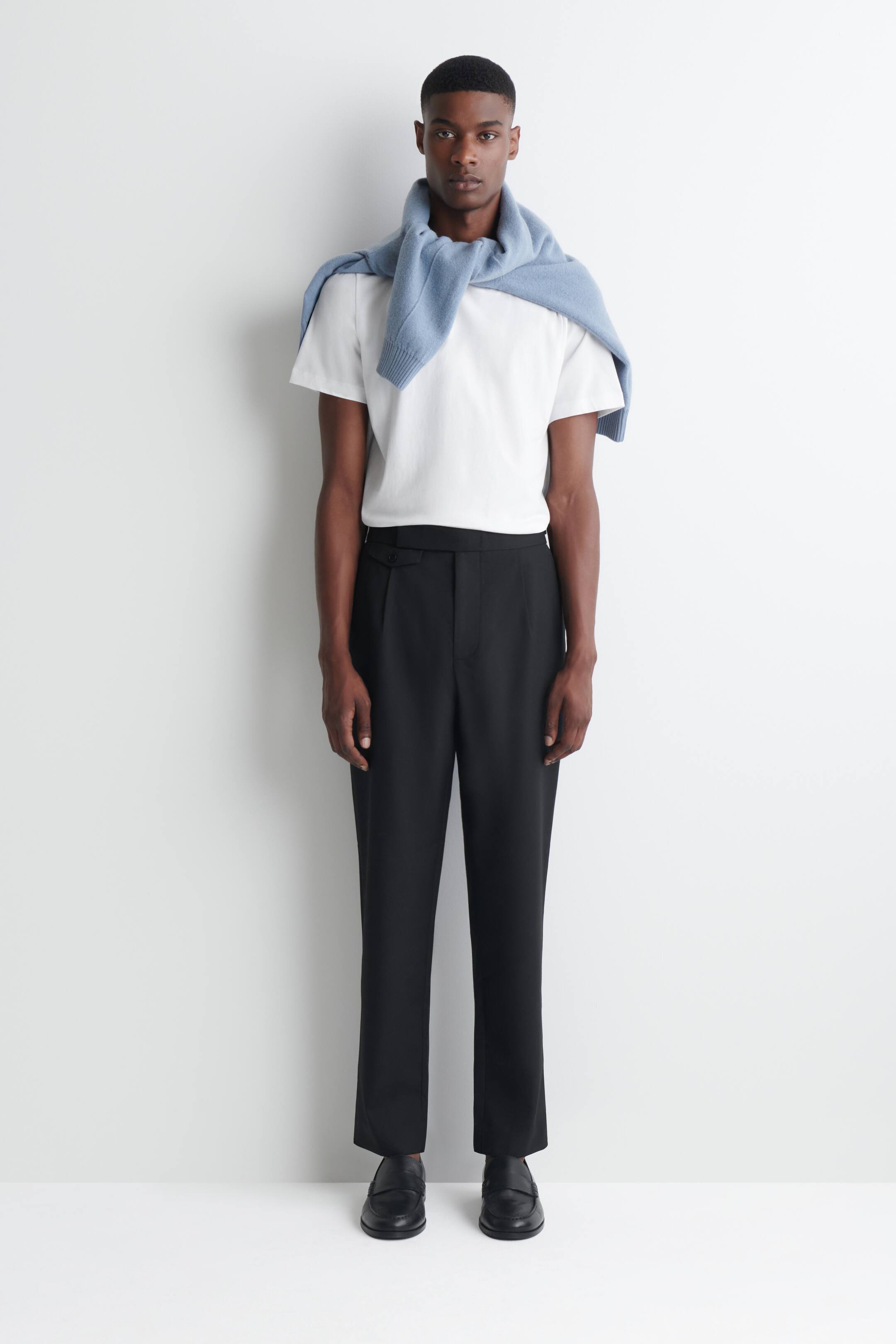 Grotere afbeelding bekijken: De model draagt een zwarte wollen slim-fit pantalon met rechte pijpen, met een gestructureerde tailleband met een tabdetail met knoop en een strakke vouw aan de voorkant. De pantalon is gestyled met een witte T-shirt die in de tailleband is ingestopt en gecombineerd met zwarte leren loafers.
