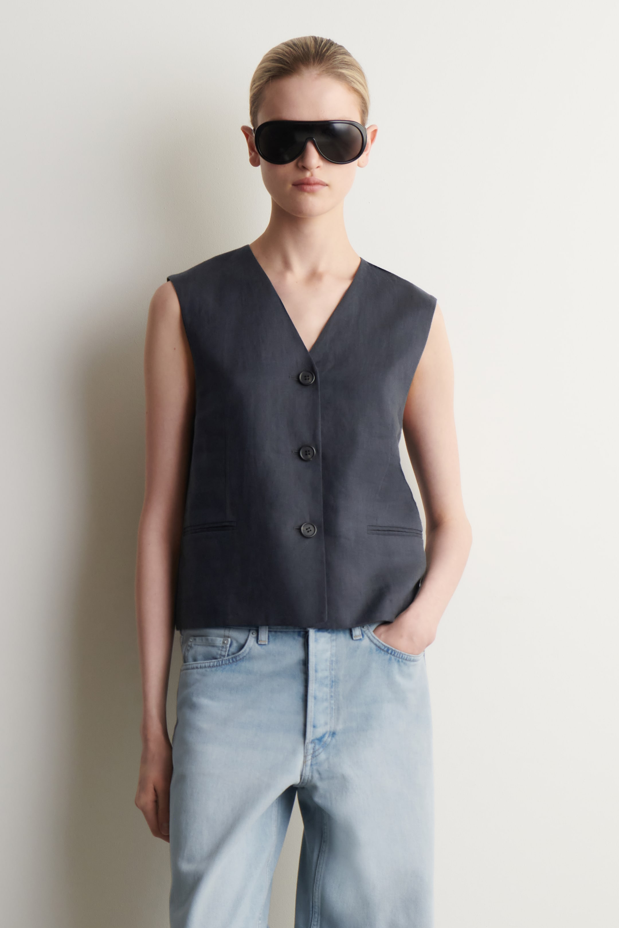 Zobrazit větší obrázek: Tmavě šedá vesta bez rukávů je prezentována zepředu, s výstřihem do V a zapínáním na tři knoflíky vpředu. Dále jsou vidět dvě horizontální lištové kapsy na spodní části přední strany.