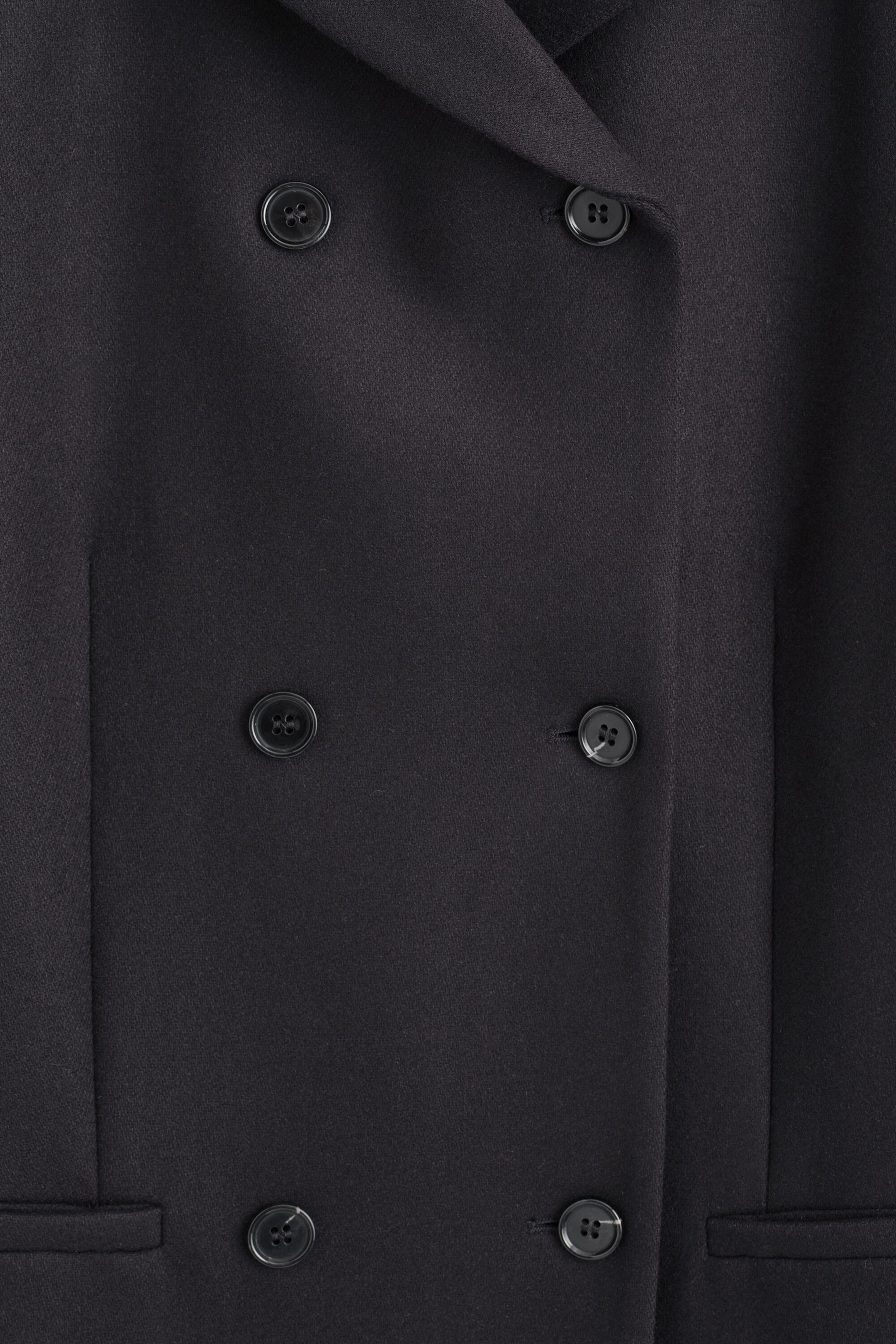 A close-up front view of a black wool-blend coat highlighting its double-breasted closure with six dark buttons. The textured fabric showcases clean vertical seams and two side welt pockets. A close-up front view of a black wool-blend coat highlighting its double-breasted closure with six dark buttons. The textured fabric showcases clean vertical seams and two side welt pockets.