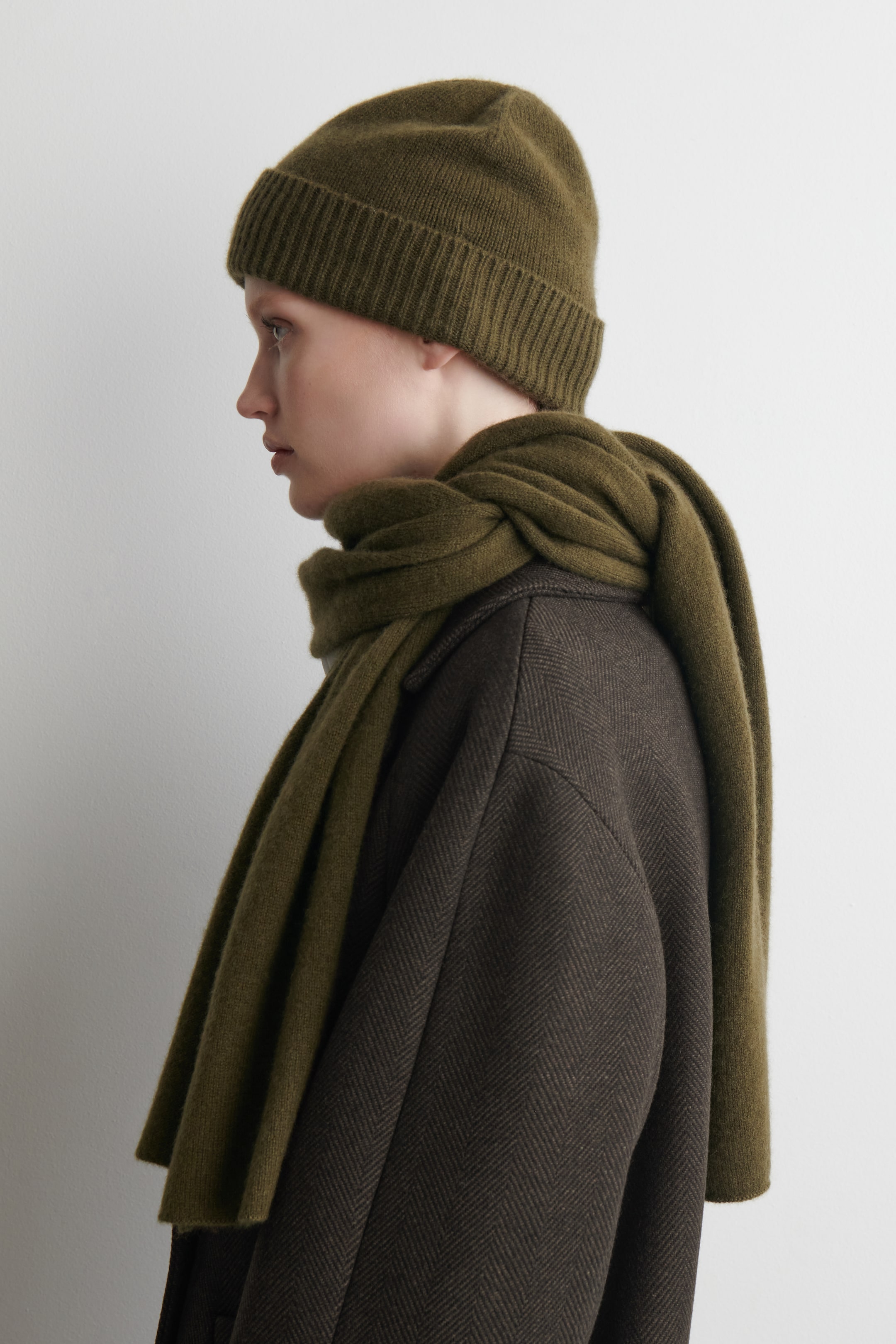 Größeres Bild anzeigen: Eine olivgrüne Strickmütze mit geripptem Rand wird getragen, aus einer Seitenansicht gesehen, während ein passender olivgrüner weicher Schal um den Hals und über die Schultern eines dunkelbraunen Fischgrätmantels drapiert wird.