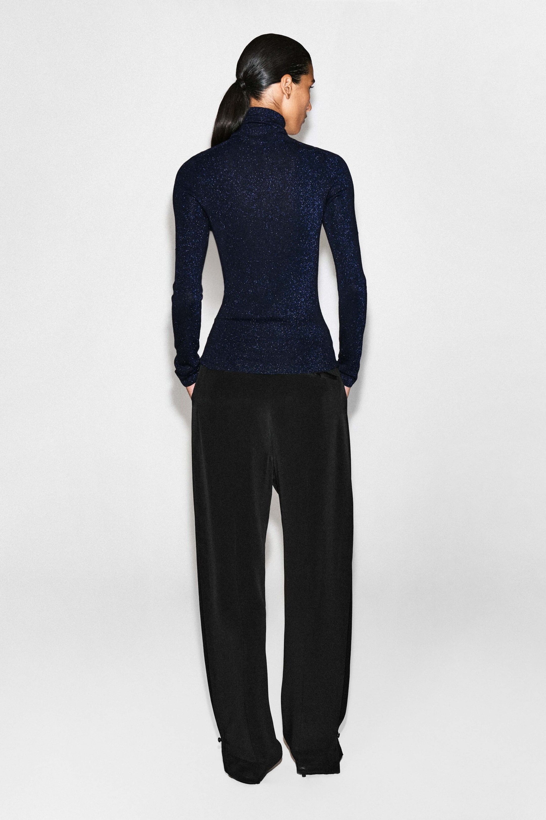 The sparkly seamless wool funnel-neck top is shown from the back, highlighting its form-fitting silhouette and shimmering dark navy knit. The long-sleeved top features a high funnel neck and a seamless design that drapes smoothly over the waistband of black trousers.
