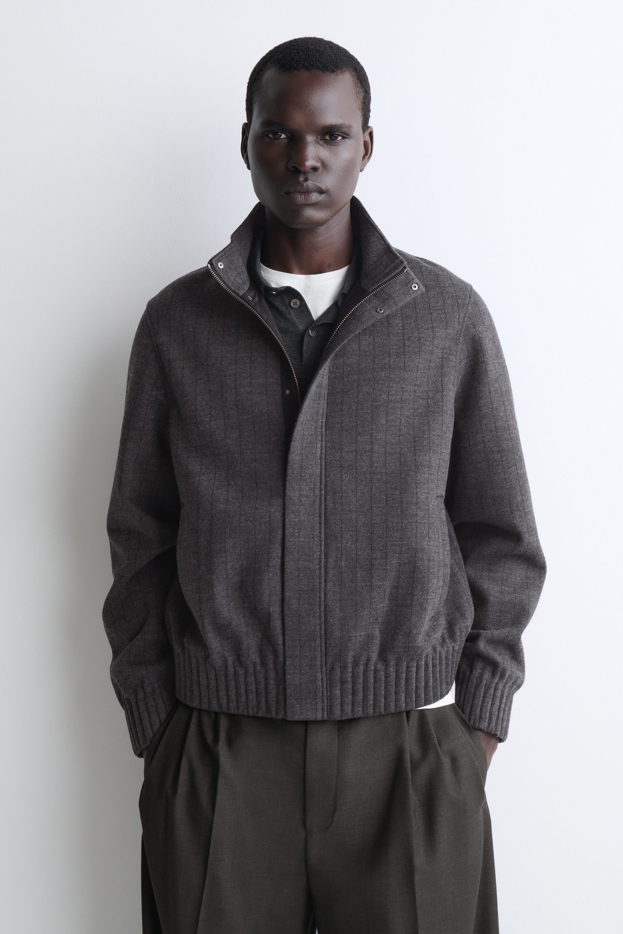 This gray bomber jacket features a vertical ribbed pattern, a high stand collar with snap-button details, and ribbed hem and cuffs, shown from the front. The jacket is worn partially unzipped, layered over a dark button-up shirt and a white T-shirt, and paired with dark pleated trousers. This gray bomber jacket features a vertical ribbed pattern, a high stand collar with snap-button details, and ribbed hem and cuffs, shown from the front. The jacket is worn partially unzipped, layered over a dark button-up shirt and a white T-shirt, and paired with dark pleated trousers.