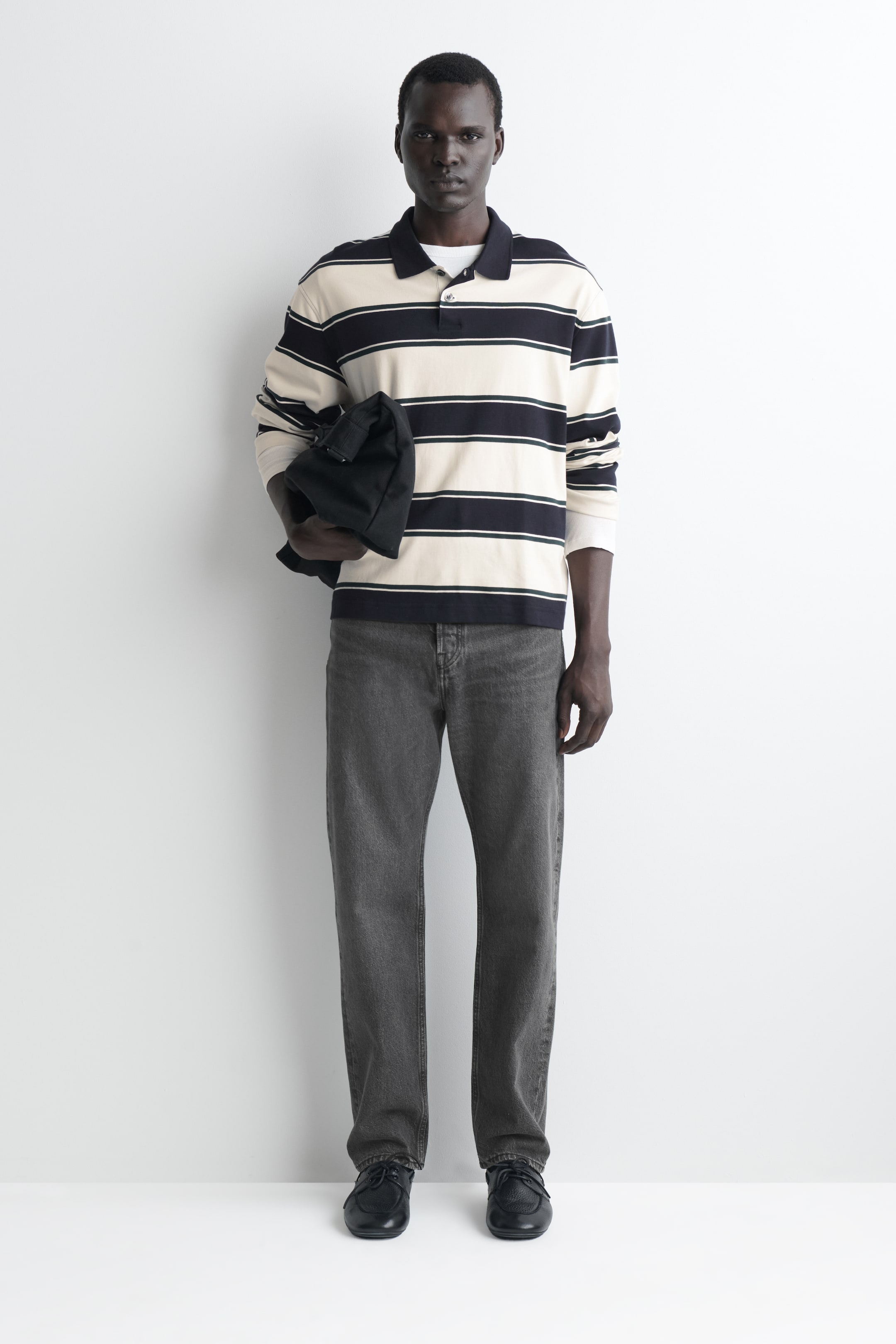 Grösseres Bild ansehen: Ein Langarm-Poloshirt mit cremefarbenen und marineblauen horizontalen Streifen wird aus der Vorderansicht gezeigt und mit gerade geschnittenen grauen Denim-Jeans kombiniert. Das Shirt verfügt über einen einfarbig marineblauen Kragen, eine Knopfleiste und hellfarbene Ärmelabschlüsse, während eine schwarze Stofftasche unter dem Arm getragen wird.