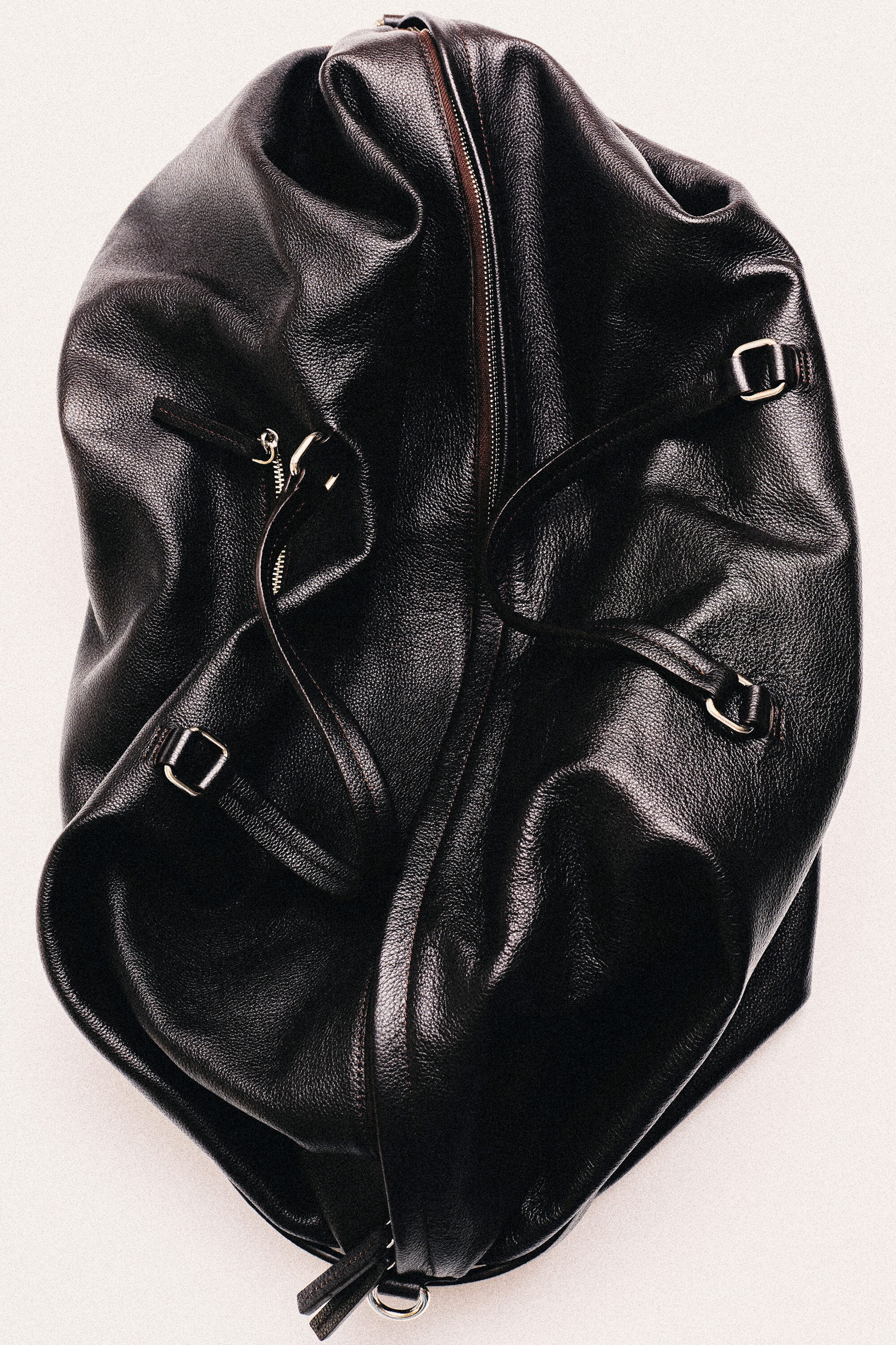The Departure Duffle Bag in black pebbled leather is resting in a slouchy, relaxed position, showcasing its soft structure from a top-down view. It features a central main zipper, top carry handles with silver-toned metal hardware, and an exterior zippered pocket.