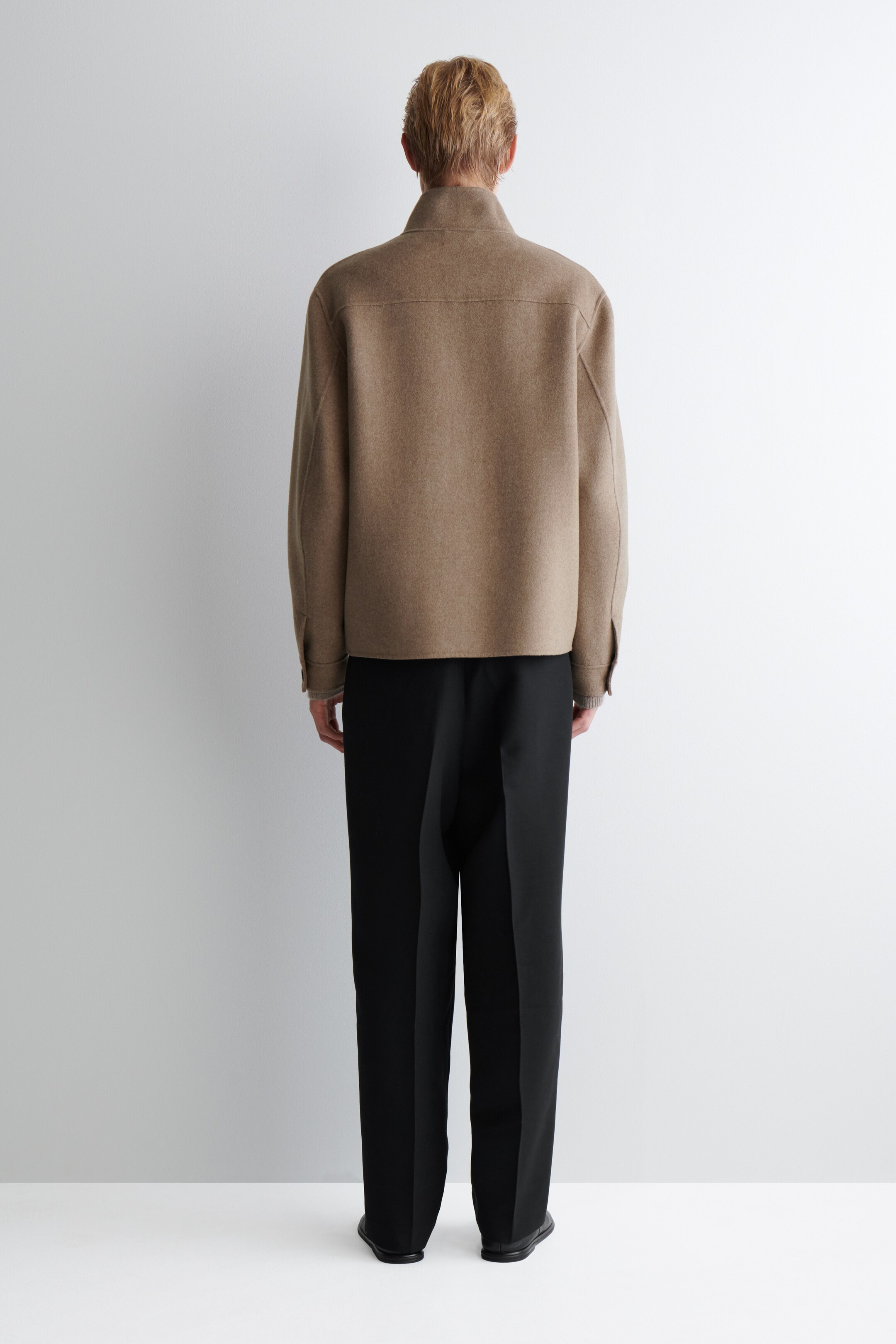 Ver imagen más grande: Se está viendo una chaqueta corta de color camel desde atrás, con un cuello levantado, una costura horizontal a lo largo de la parte superior de la espalda y puños con botones. Se están usando pantalones negros de pierna ancha debajo de la chaqueta, con una prenda de color claro visible debajo de los puños de la chaqueta.