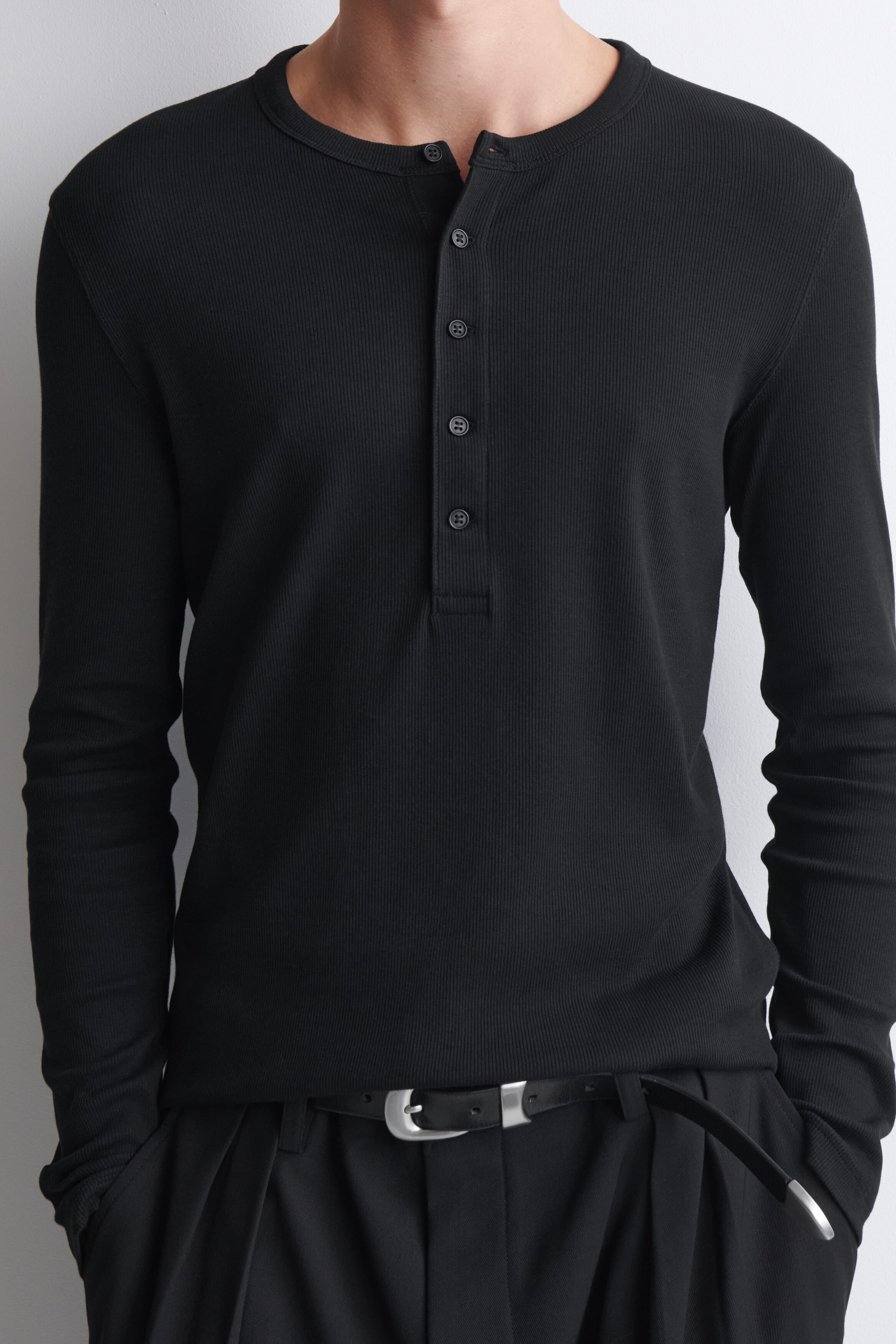 The slim ribbed cotton long-sleeved henley T-shirt is shown in a form-fitting silhouette with a fine ribbed texture in a front close-up view. The black top features a round neckline with a five-button placket and is styled tucked into black trousers with a silver-buckled belt. The slim ribbed cotton long-sleeved henley T-shirt is shown in a form-fitting silhouette with a fine ribbed texture in a front close-up view. The black top features a round neckline with a five-button placket and is styled tucked into black trousers with a silver-buckled belt.