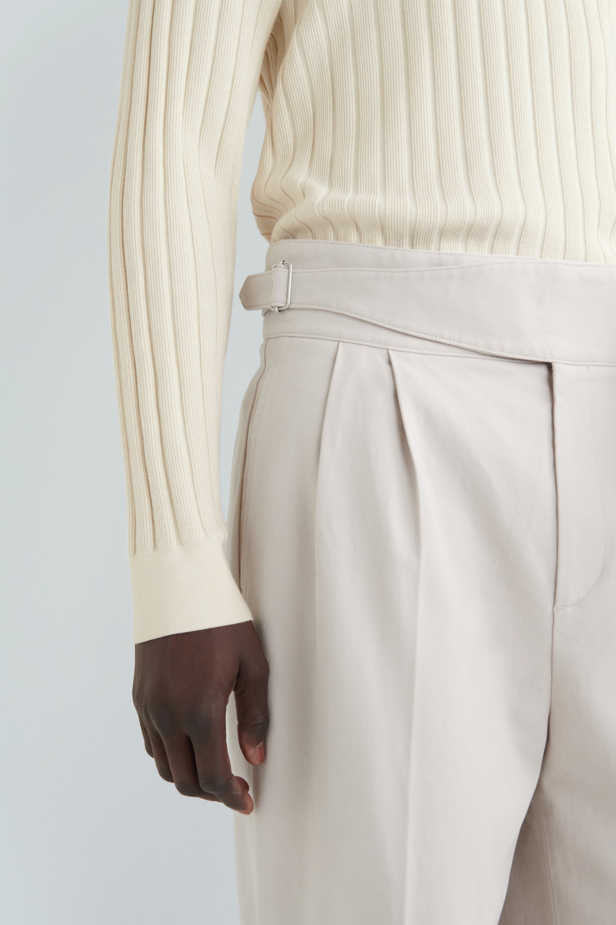 Agrandir l'image: Un pantalon beige clair est doté de plis doubles à l'avant et de réglages latéraux avec une boucle de ton argenté. Un pull en maille côtelée blanc cassé, avec des poignets en côtes visibles, est porté rentré dans le pantalon.