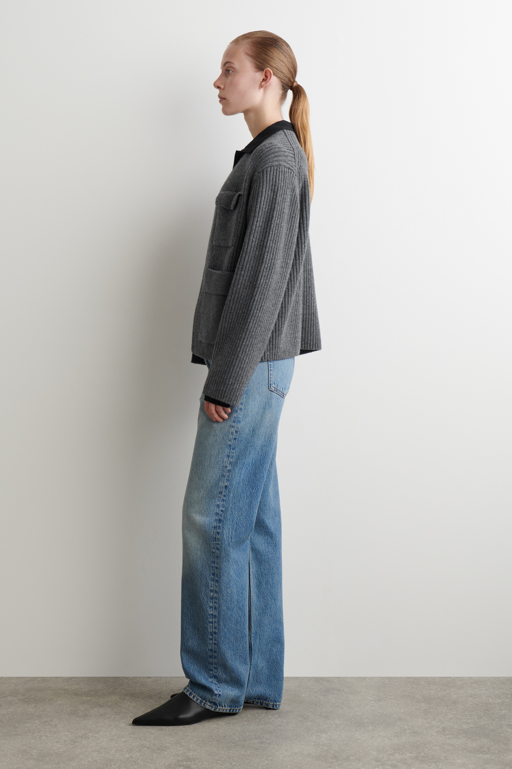Visualizza immagine più grande: Una giacca a maglia grigia a coste è presentata in un profilo laterale, con due tasche applicate frontali. È indossata sopra un top nero con colletto, abbinata a jeans a gamba larga a lavaggio chiaro con una cucitura centrale anteriore visibile, e scarpe basse a punta nere.