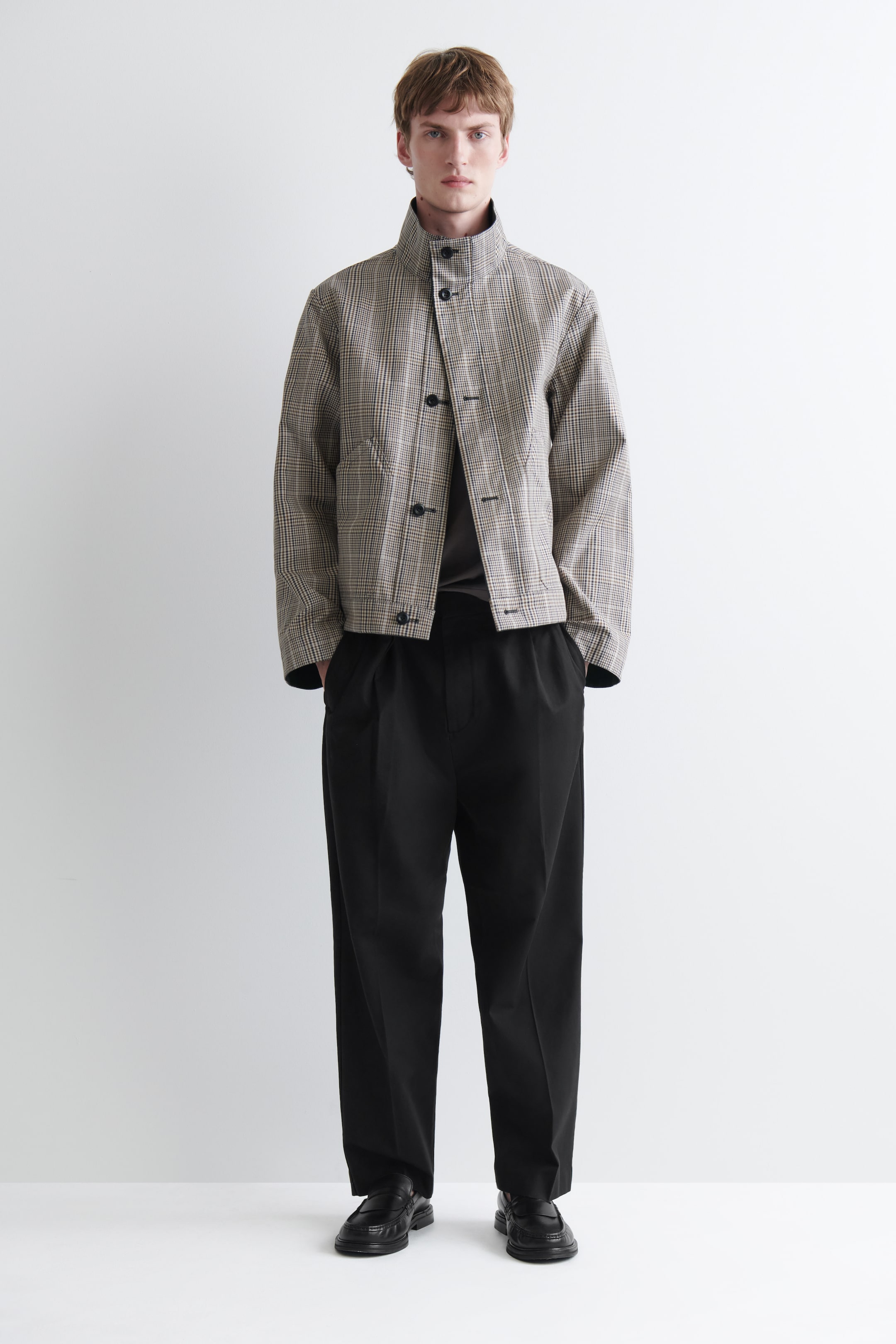 Grösseres Bild ansehen: Eine beige und braun karierte Jacke wird präsentiert, mit hochgeschlossenem, geknöpftem Kragen und einem vorne teilweise aufgeknöpften Verschluss mit drei sichtbaren dunklen Knöpfen. Auf der Vorderseite befinden sich zwei schräge Leistentaschen, darunter wird ein dunkles T-Shirt getragen.
