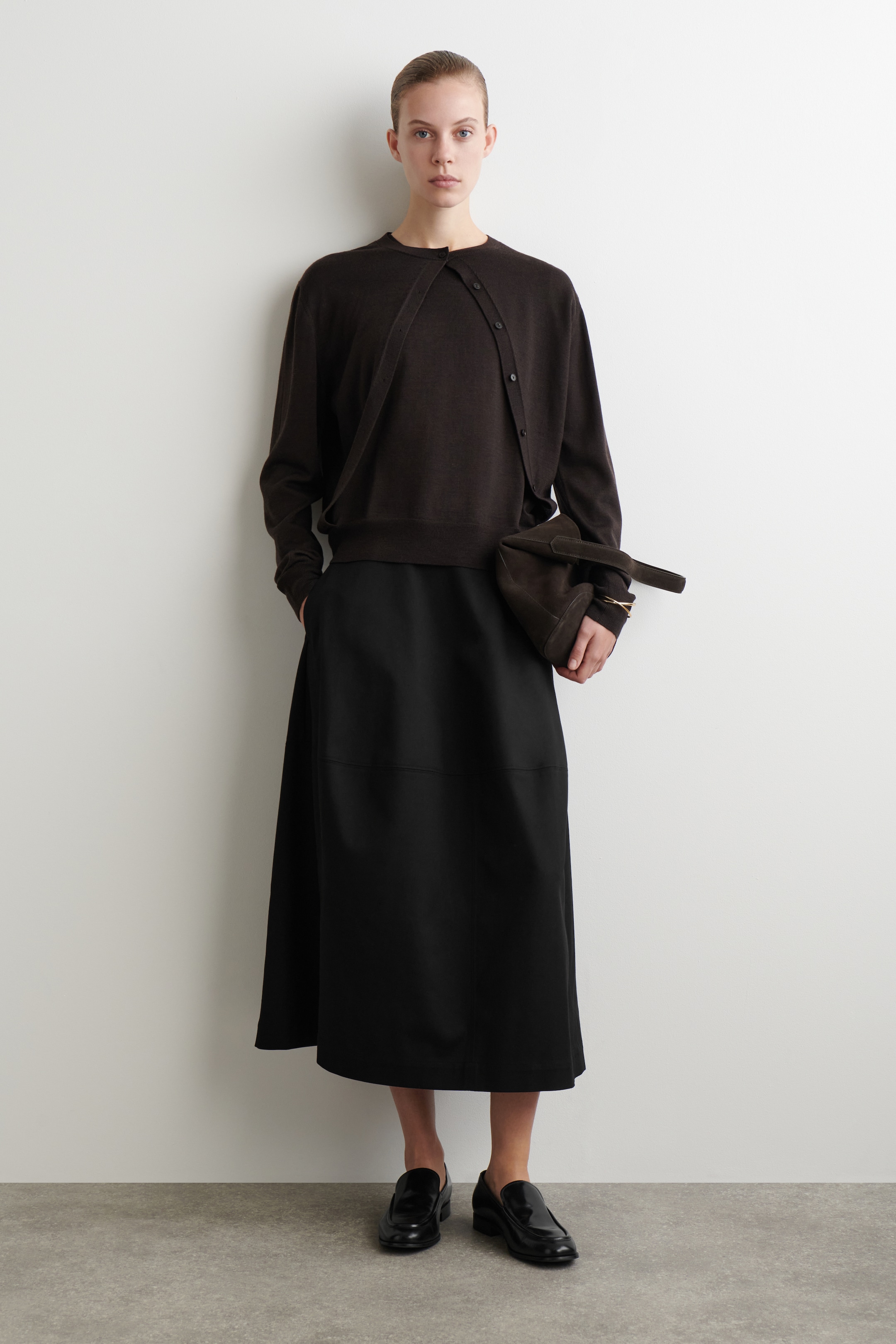 Ingrandisci l'immagine: Un top a maglia marrone scuro con scollo rotondo è indossato, stratificato con un cardigan marrone scuro abbinato che è drappeggiato diagonalmente sul davanti e fissato con bottoni a destra. Questo ensemble è completato con una gonna nera a linea A di lunghezza midi, che presenta una cucitura orizzontale, mocassini neri lucidi e una borsa strutturata marrone scuro tenuta in mano.