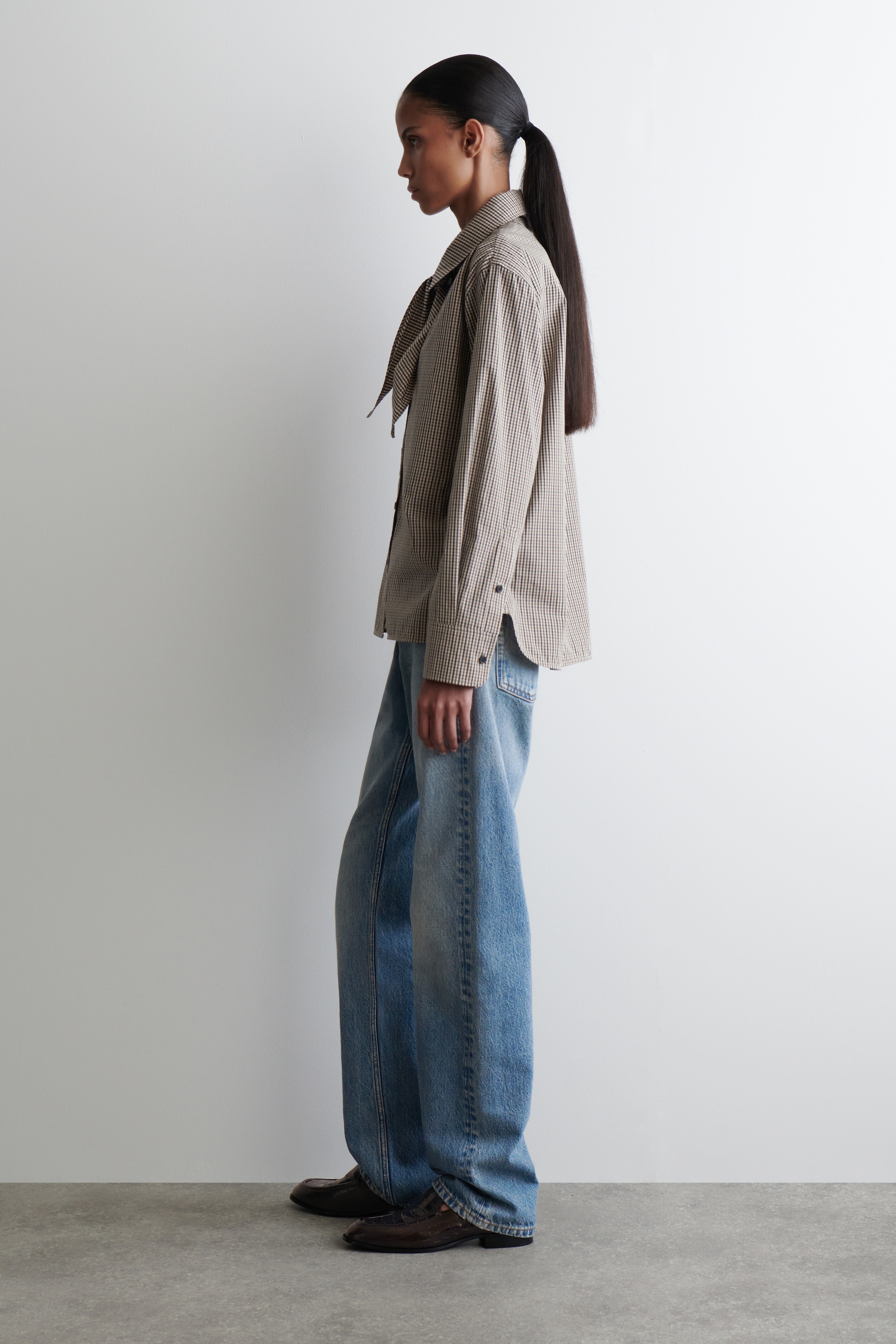 Agrandir l'image: Une chemise à carreaux ample en marron et blanc cassé est portée, avec un col pointu et un détail de cravate pendante, et des manches longues avec des poignets boutonnés. Elle est stylée par-dessus un jean bleu à jambes larges et délavage clair et des mocassins marron foncé, montrés de profil.