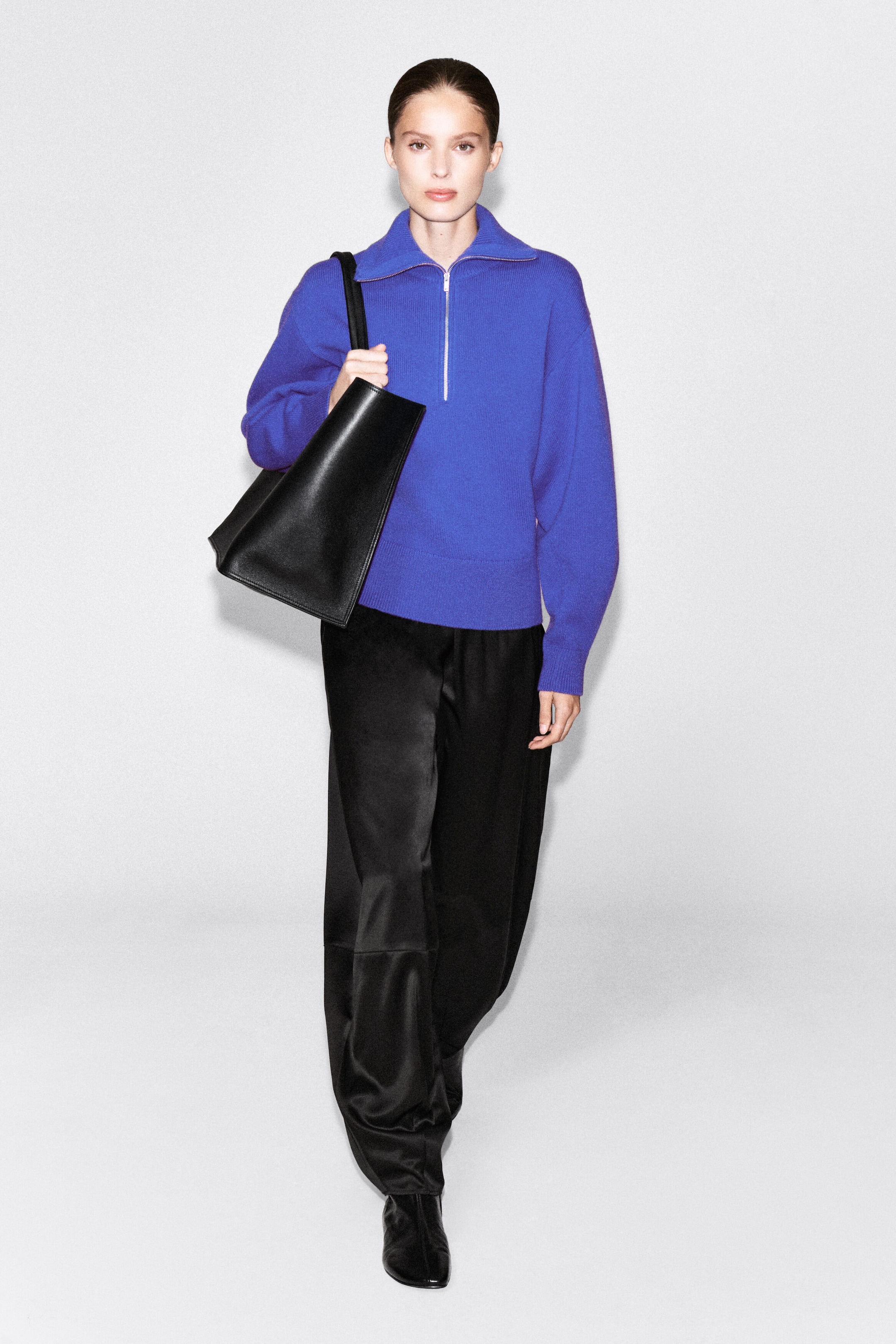 A bright blue knitted sweater featuring a half-zip funnel neck and relaxed, sculptural sleeves is being worn by the model. The sweater is styled with black trousers and a black leather shoulder bag. A bright blue knitted sweater featuring a half-zip funnel neck and relaxed, sculptural sleeves is being worn by the model. The sweater is styled with black trousers and a black leather shoulder bag.