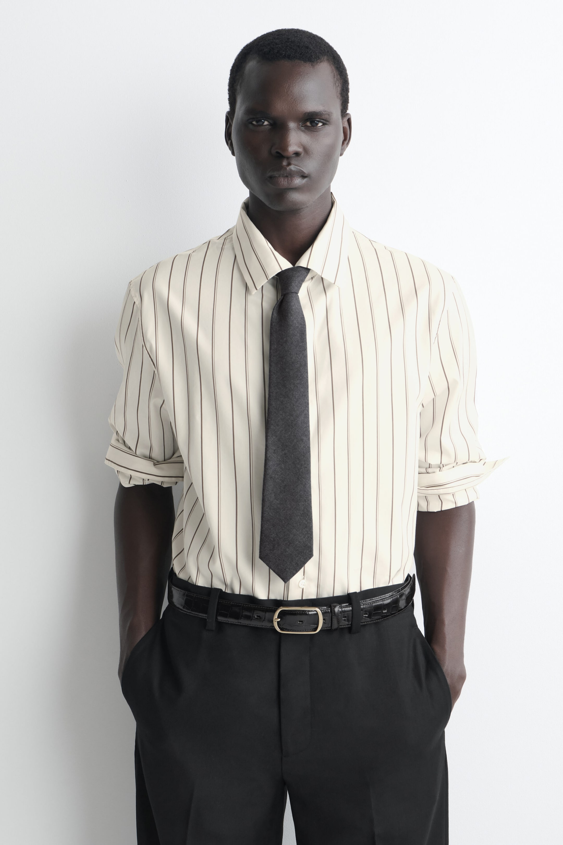 A cream-coloured dress shirt featuring thin, dark brown vertical stripes is shown from the front, designed with a classic collar and sleeves rolled up to the elbows. The shirt is tucked into black trousers secured with a black croc-effect belt and paired with a solid dark grey tie. A cream-coloured dress shirt featuring thin, dark brown vertical stripes is shown from the front, designed with a classic collar and sleeves rolled up to the elbows. The shirt is tucked into black trousers secured with a black croc-effect belt and paired with a solid dark grey tie.