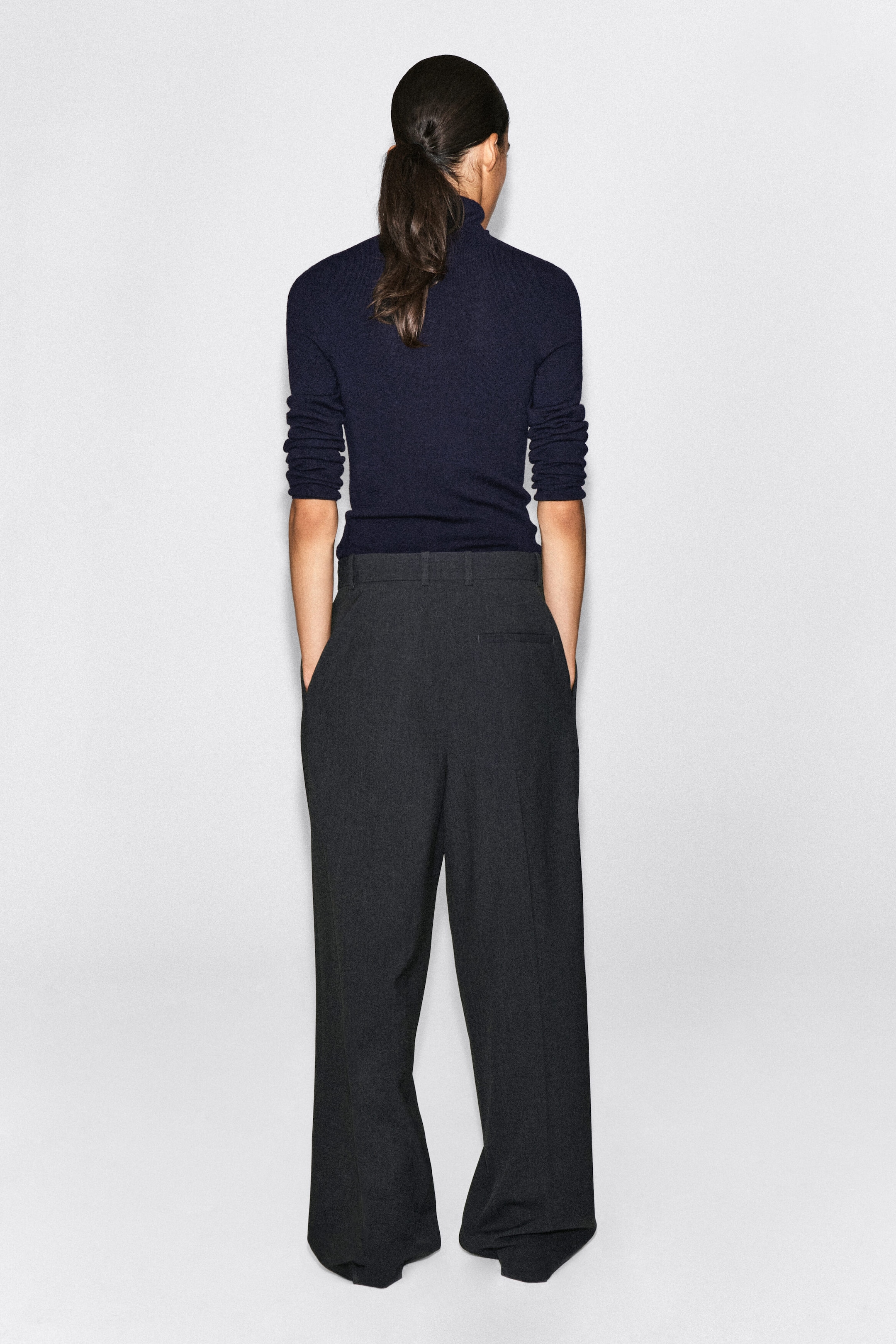 A pair of dark grey, wide-leg trousers is shown from the back, featuring a high-waisted design with belt loops and a single back welt pocket. The trousers are styled with a tucked-in navy blue roll-neck sweater, emphasising their relaxed fit and structured drape. A pair of dark grey, wide-leg trousers is shown from the back, featuring a high-waisted design with belt loops and a single back welt pocket. The trousers are styled with a tucked-in navy blue roll-neck sweater, emphasising their relaxed fit and structured drape.