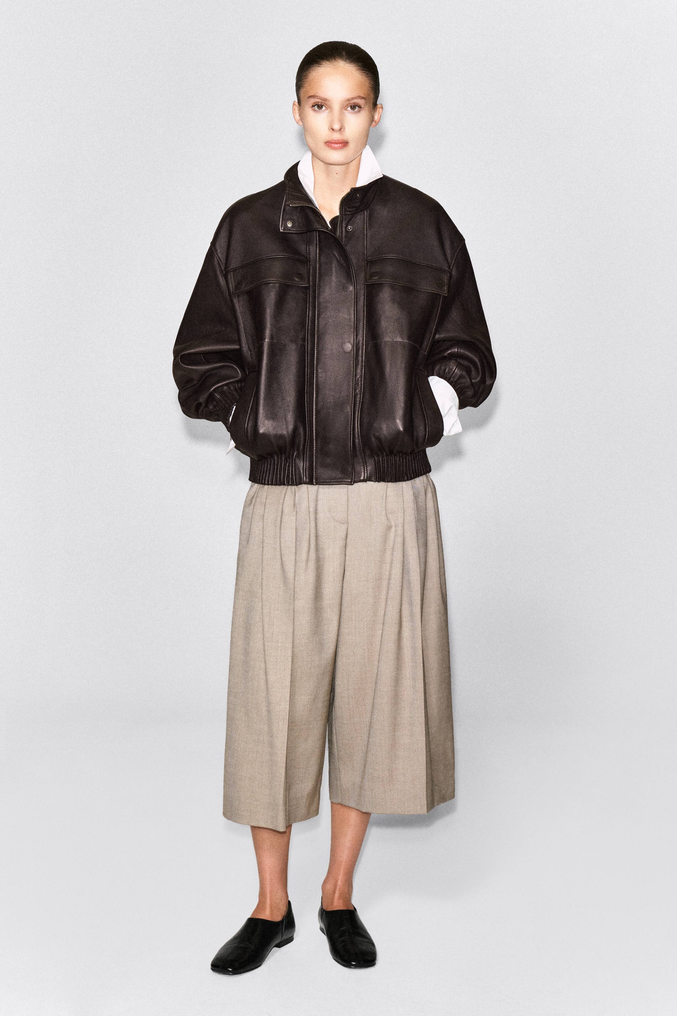 Ver imagen más grande: La modelo lleva unos culottes de mezcla de lana plisados en un tono beige claro, con una silueta cropped de pierna ancha y profundos pliegues delanteros. Los culottes se combinan con una chaqueta de cuero marrón oscuro, una camisa blanca con cuello y zapatos negros de cuero tipo slip-on.