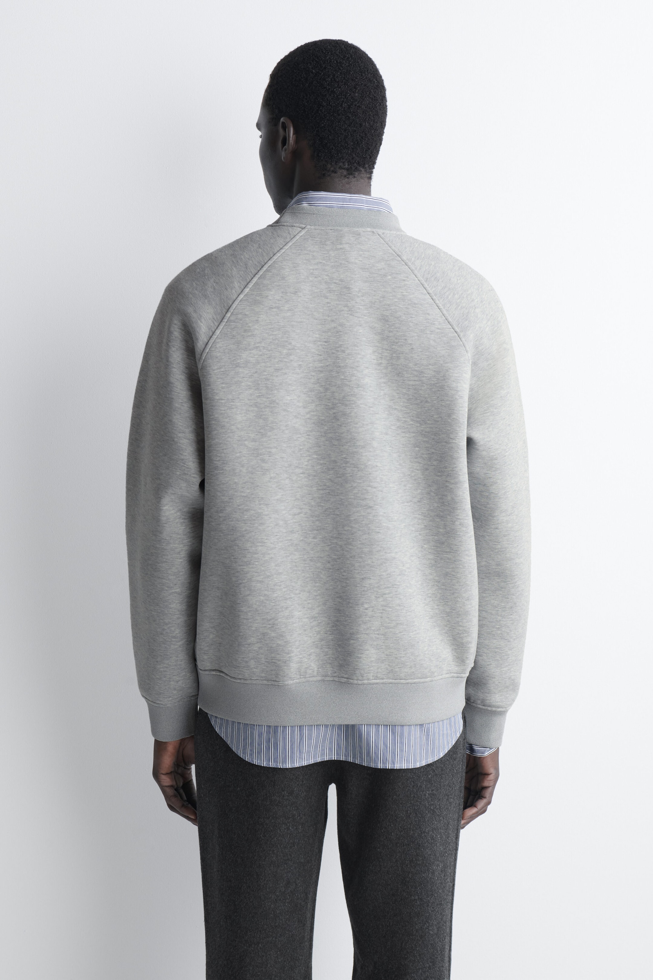 The light grey relaxed scuba sweatshirt is shown from the back, featuring raglan sleeves, a ribbed collar, and a ribbed hem and cuffs. It is layered over a blue-and-white striped shirt that is visible at the neckline and extends below the hem. The light grey relaxed scuba sweatshirt is shown from the back, featuring raglan sleeves, a ribbed collar, and a ribbed hem and cuffs. It is layered over a blue-and-white striped shirt that is visible at the neckline and extends below the hem.
