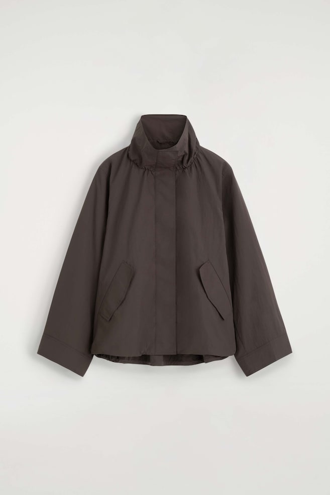 Die Nylon-Jacke mit Funnel-Neck präsentiert sich von vorne in einer kastigen Silhouette in dunklem Braun und verfügt über einen hohen, gerafften Funnel-Neck sowie eine verdeckte vordere Knopfleiste. Zu sehen sind weite, lange Ãrmel mit genähten Manschetten, schräge Pattentaschen und ein leicht geraffter Saum. - 1