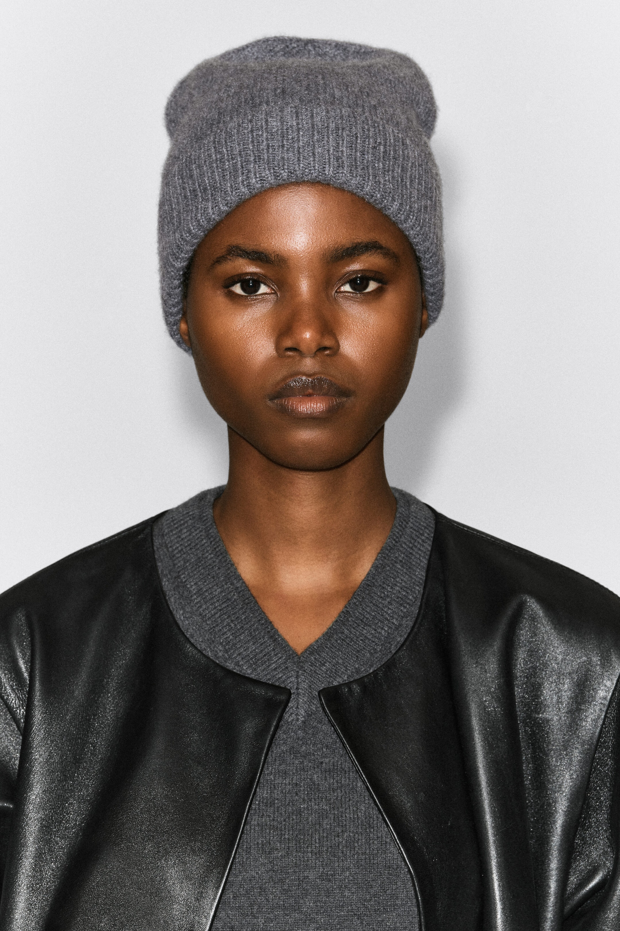 A grey ribbed knit beanie is worn on the model’s head, paired with a matching grey V-neck knit top. A black leather jacket is layered over the knit top, completing the look. A grey ribbed knit beanie is worn on the model’s head, paired with a matching grey V-neck knit top. A black leather jacket is layered over the knit top, completing the look.