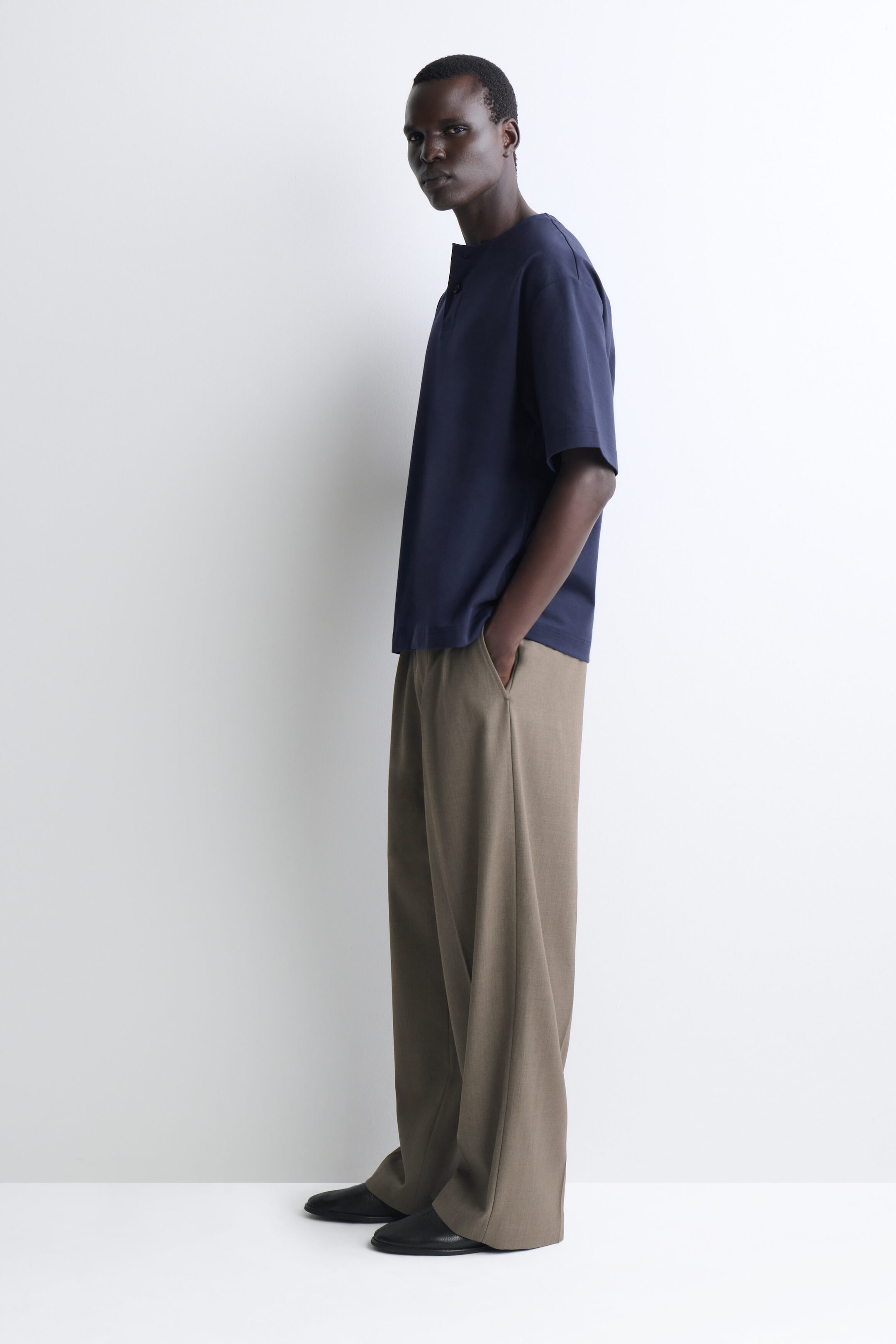 Le t-shirt henley oversize en jersey de coton est porté de profil gauche, mettant en valeur sa coupe décontractée, ses manches courtes amples et son col partiellement boutonné. Le t-shirt tombe souplement sur un pantalon à jambe large, soulignant son ourlet droit et sa coupe décontractée.