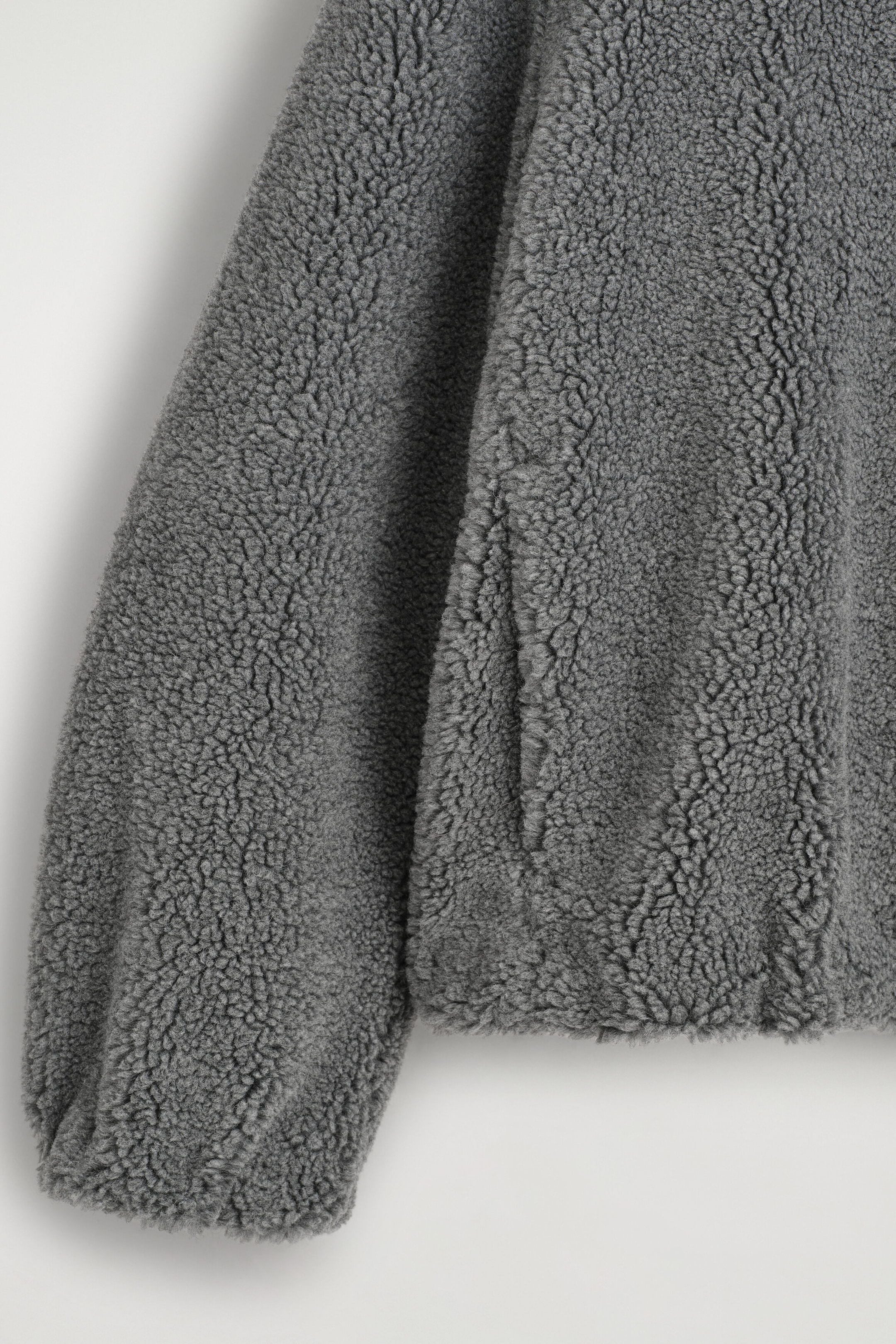 A grey teddy jacket is shown in close-up, highlighting its dense, curly fleece texture. The view focuses on the lower section, showing the sleeve, hem, and a subtle side pocket seam. A grey teddy jacket is shown in close-up, highlighting its dense, curly fleece texture. The view focuses on the lower section, showing the sleeve, hem, and a subtle side pocket seam.