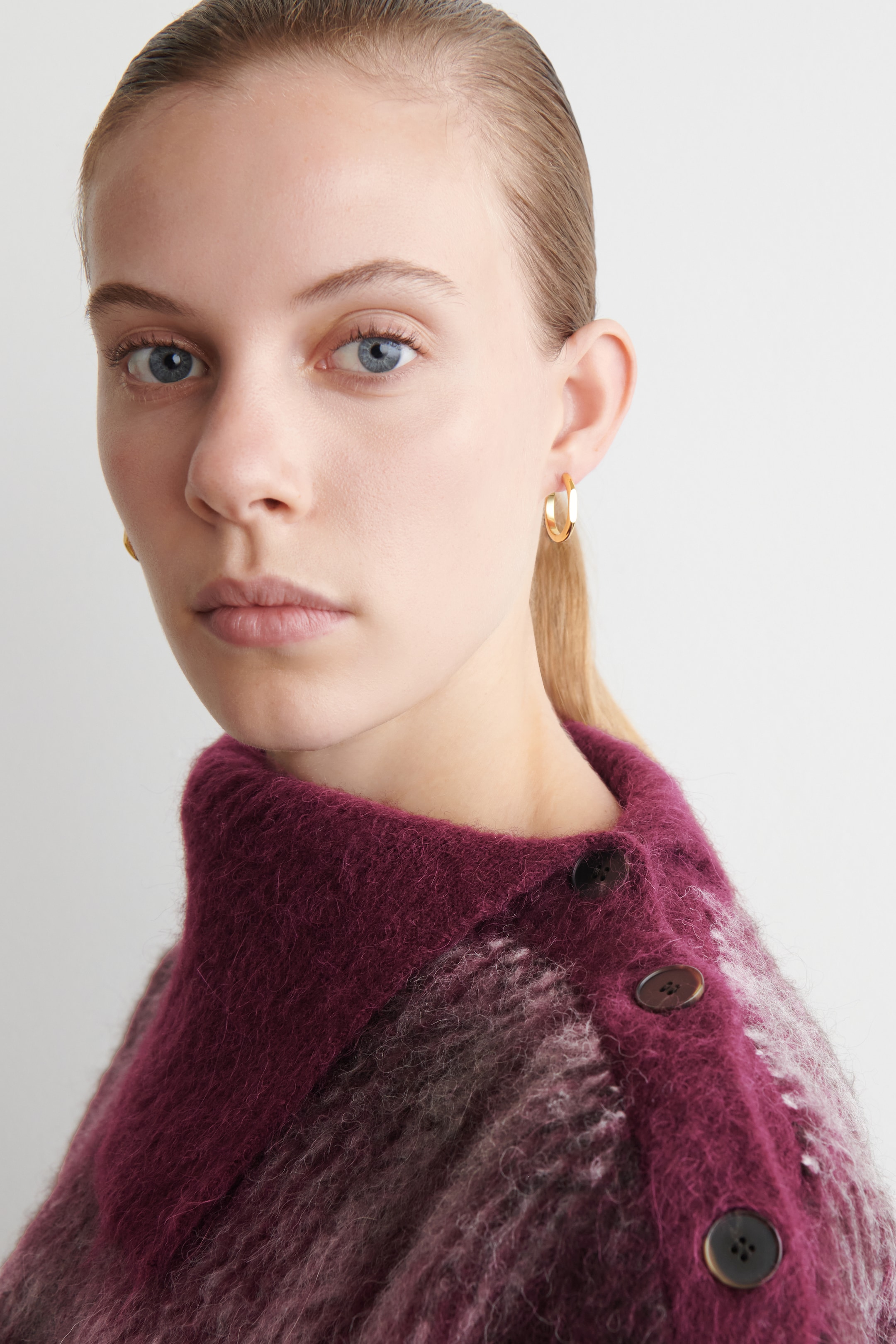 Agrandir l'image: Un pull en tricot duveteux est porté, mettant en valeur un motif varié en magenta profond et violet. Il est conçu avec un col montant haut et trois boutons ronds et sombres visibles sur l'épaule droite. Une boucle d'oreille en forme de cerceau aplati de ton doré est également portée à l'oreille droite du porteur.