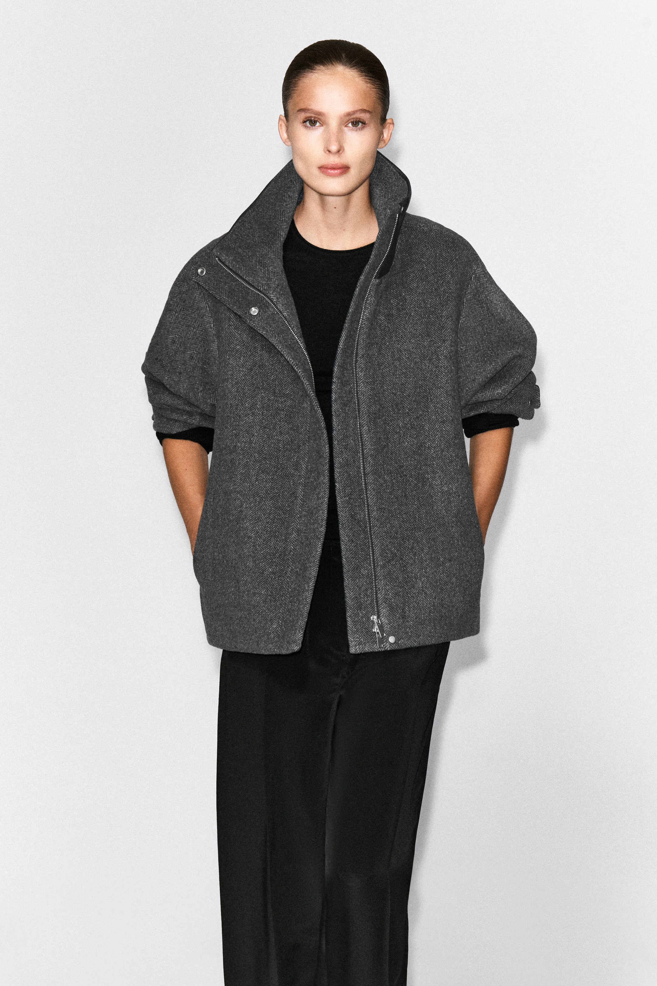 Ein grauer Wollblazer im Fischgrätenmuster mit Oversize-Silhouette wird vom Model getragen, das mit den Händen in den Seitentaschen steht. Der Blazer verfügt über einen hohen Stehkragen mit asymmetrischem Reissverschluss und ist über einem schwarzen Oberteil und einer schwarzen Hose kombiniert. Ein grauer Wollblazer im Fischgrätenmuster mit Oversize-Silhouette wird vom Model getragen, das mit den Händen in den Seitentaschen steht. Der Blazer verfügt über einen hohen Stehkragen mit asymmetrischem Reissverschluss und ist über einem schwarzen Oberteil und einer schwarzen Hose kombiniert.