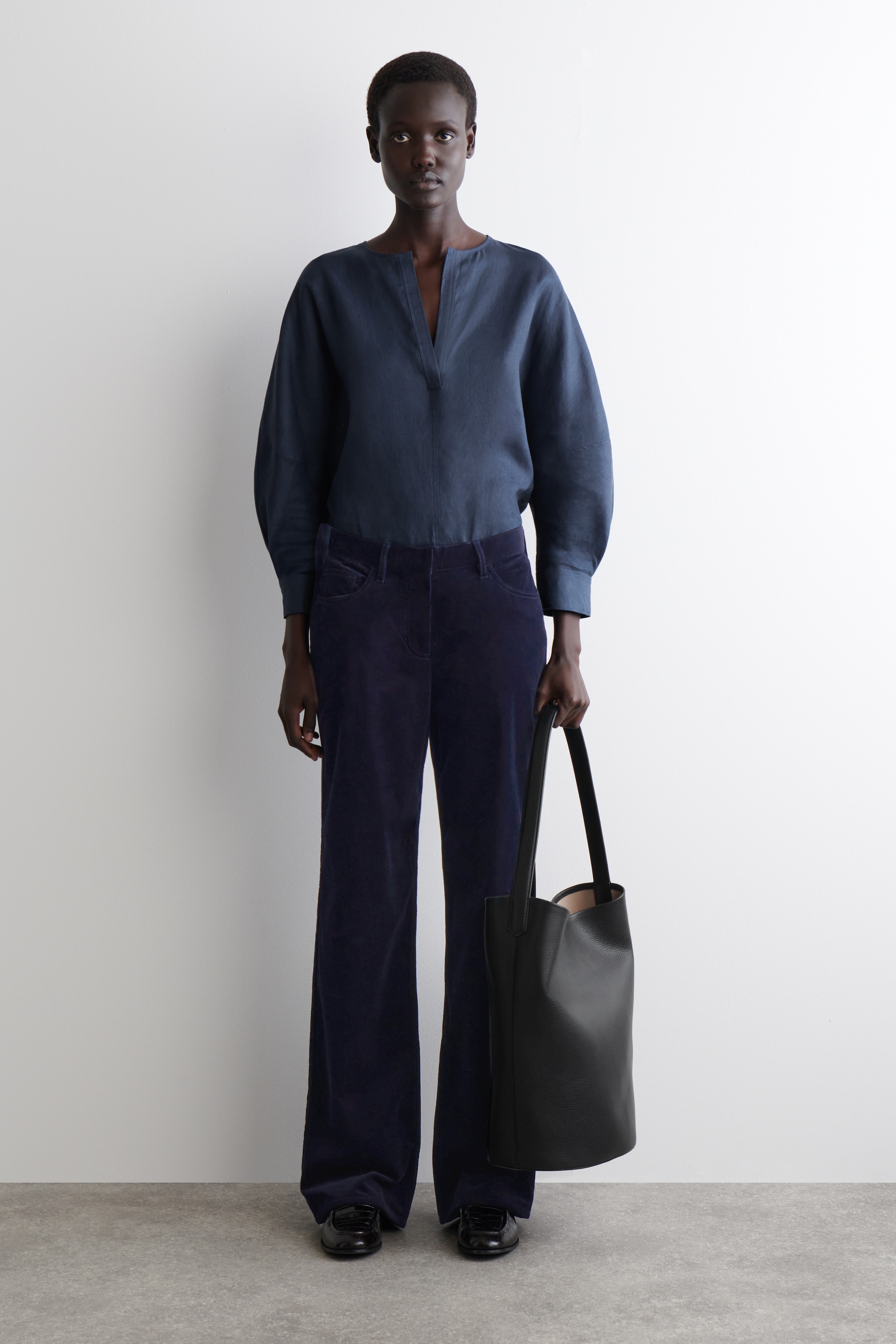 Visualizza immagine più grande: Una maglia popover a maniche lunghe blu navy con scollo a V è presentata in una vista frontale, abbinata a pantaloni a gamba larga in velluto a coste blu navy. Una borsa tote in pelle nera con texture è tenuta per il suo manico, e vengono indossate scarpe piatte nere.