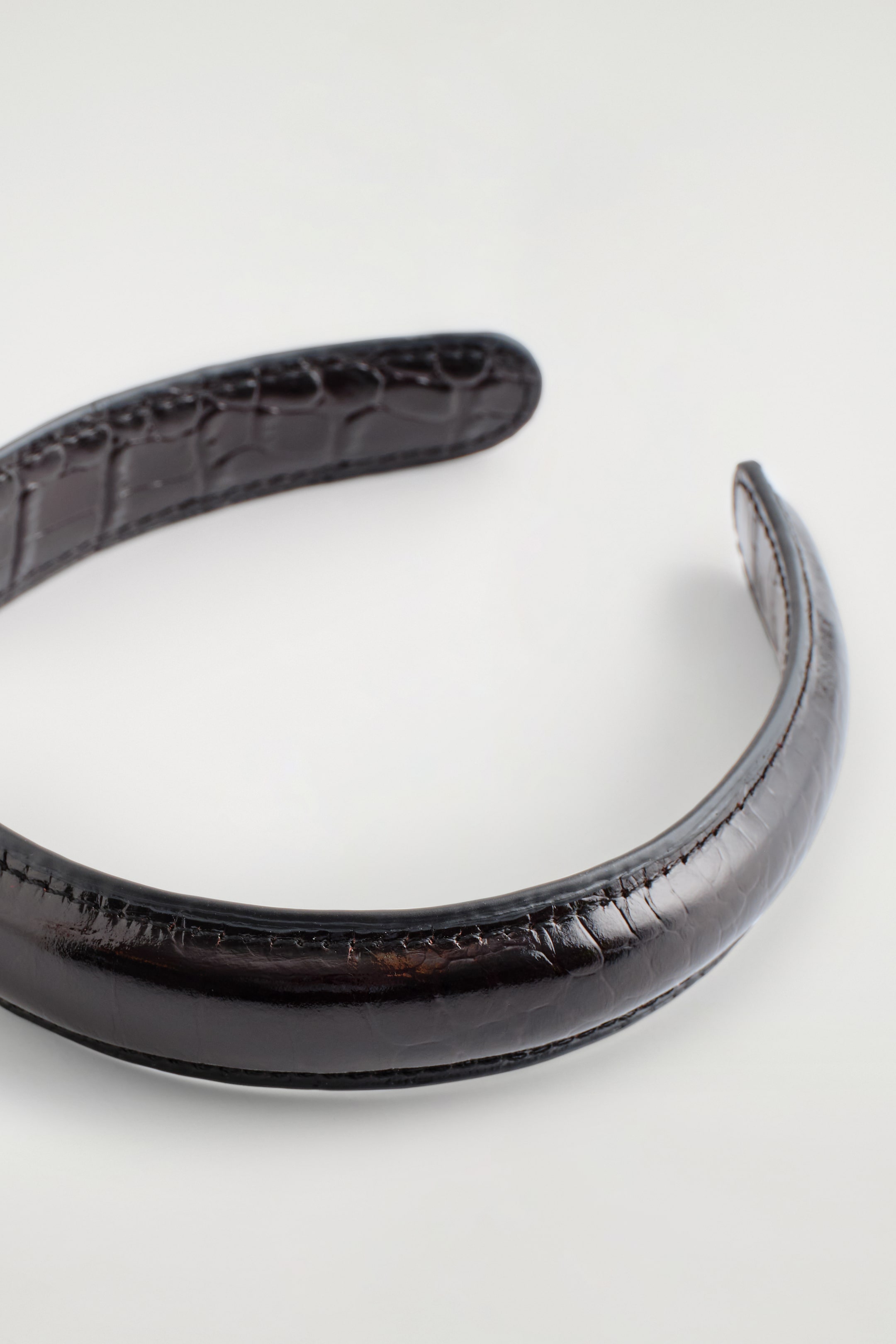 Ver imagen más grande: Una diadema de cuero marrón oscuro presenta una textura en relieve con efecto de cocodrilo. Está curvada, con costuras visibles a lo largo de sus bordes, y se presenta desde un ángulo inferior, mostrando un brillo sutil.