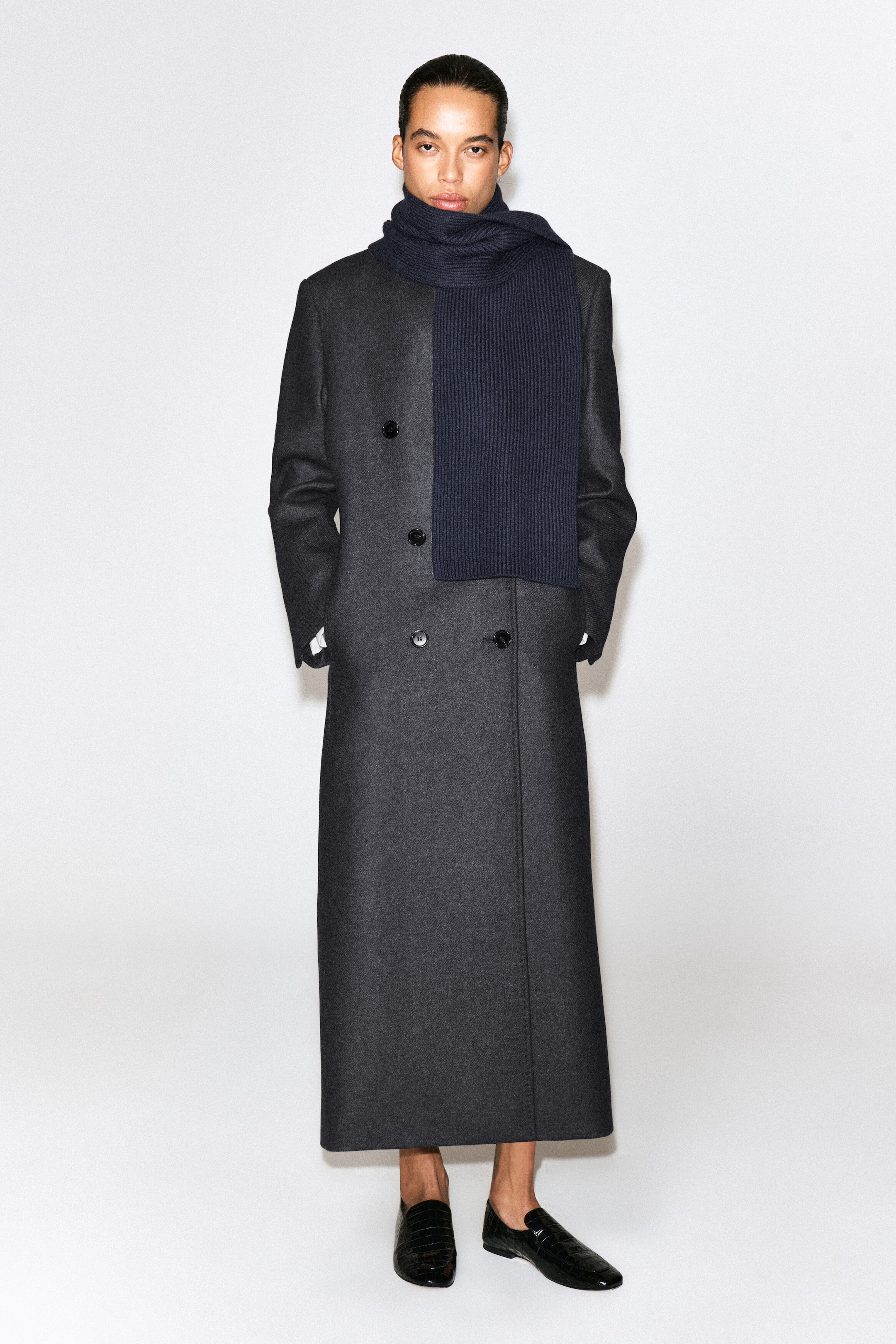 A chunky ribbed wool scarf in dark navy is being worn wrapped around the neck, with one end draping straight down the front over a long, dark grey double-breasted coat. The front-facing view showcases the scarf's thick, textured rib-knit structure and snug fit. A chunky ribbed wool scarf in dark navy is being worn wrapped around the neck, with one end draping straight down the front over a long, dark grey double-breasted coat. The front-facing view showcases the scarf's thick, textured rib-knit structure and snug fit.