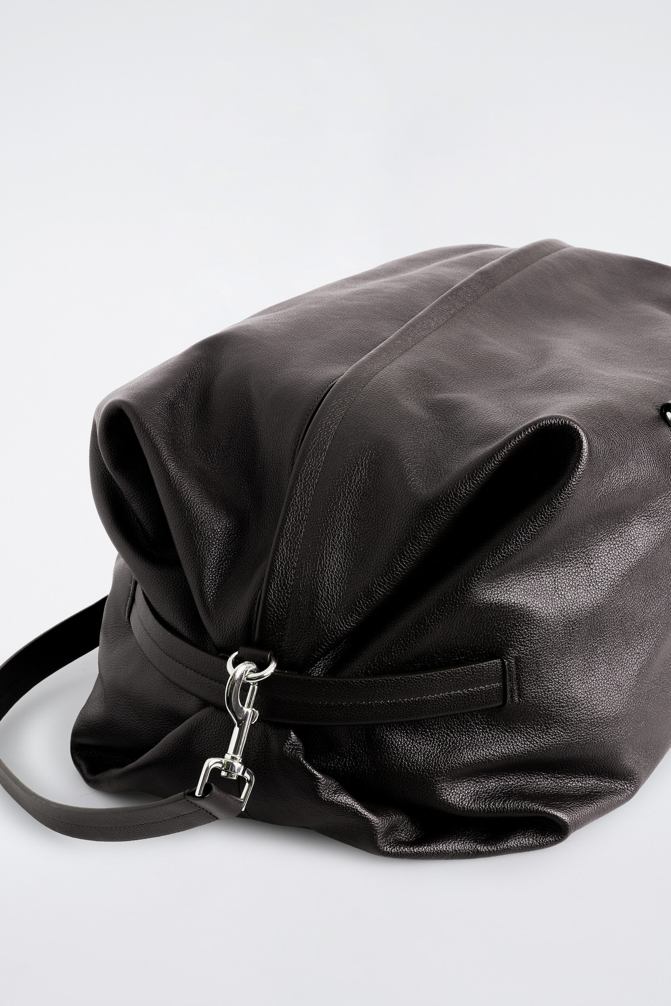 The Departure Duffle Bag in dark brown leather is resting on its side, showcasing its soft, slouchy structure and textured pebbled finish. A silver-tone metal lobster clasp is securing a matching leather shoulder strap to a loop on the folded side panel.