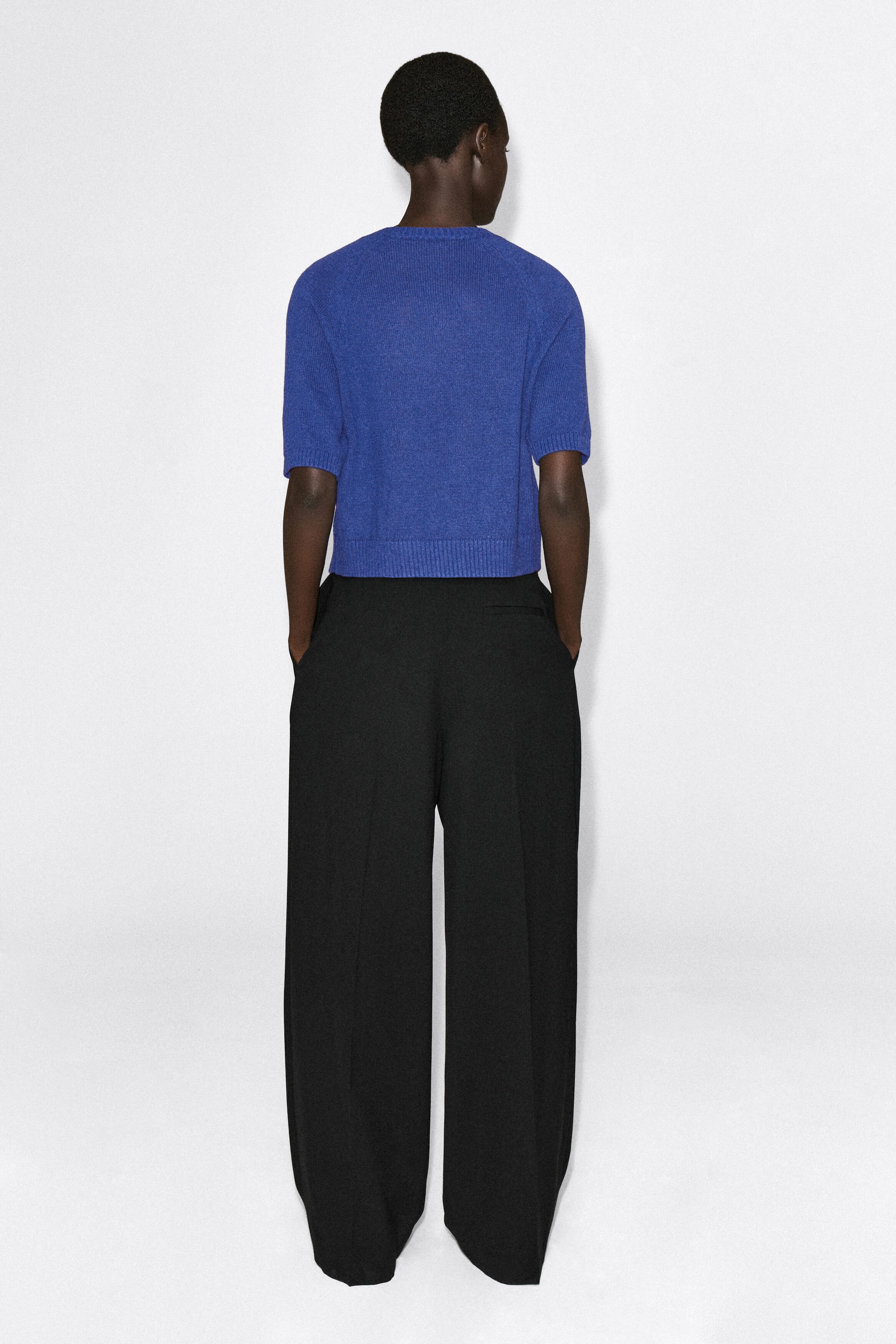 The Shrunken Knitted Linen T-shirt is shown from the back, highlighting its bright blue knit texture, cropped length, and ribbed detailing at the hem and cuffs. It is paired with wide-leg black trousers to showcase its shrunken fit and structured silhouette.