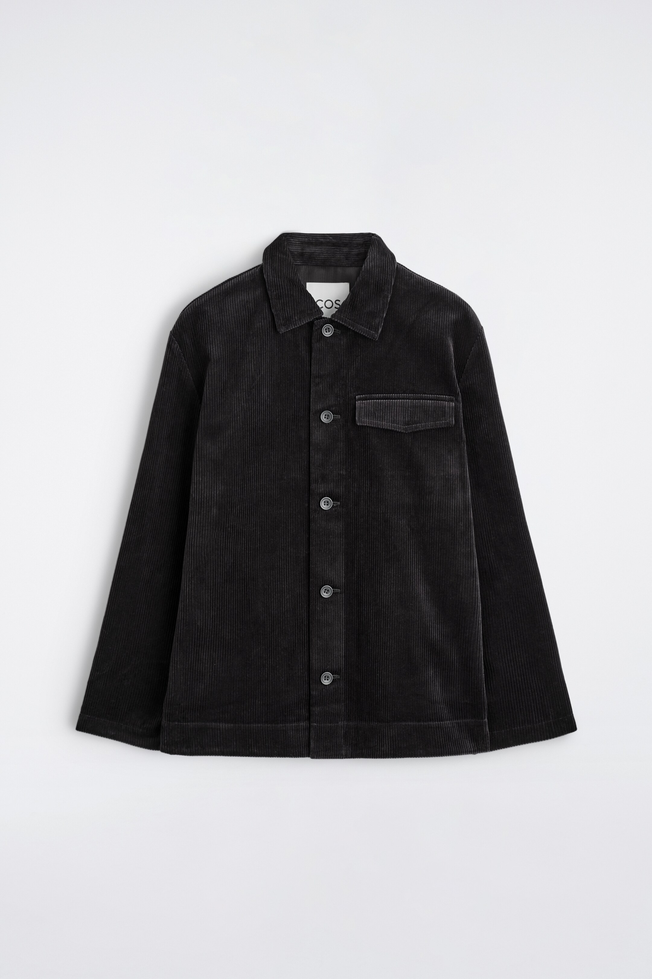 View larger image: A black corduroy overshirt is presented flat, facing FRONT, featuring a pointed collar and a button-down front closure with tonal buttons. A single flap pocket is visible on the left chest, and the overshirt has a relaxed fit with long sleeves, showing visible vertical wales in the fabric.
