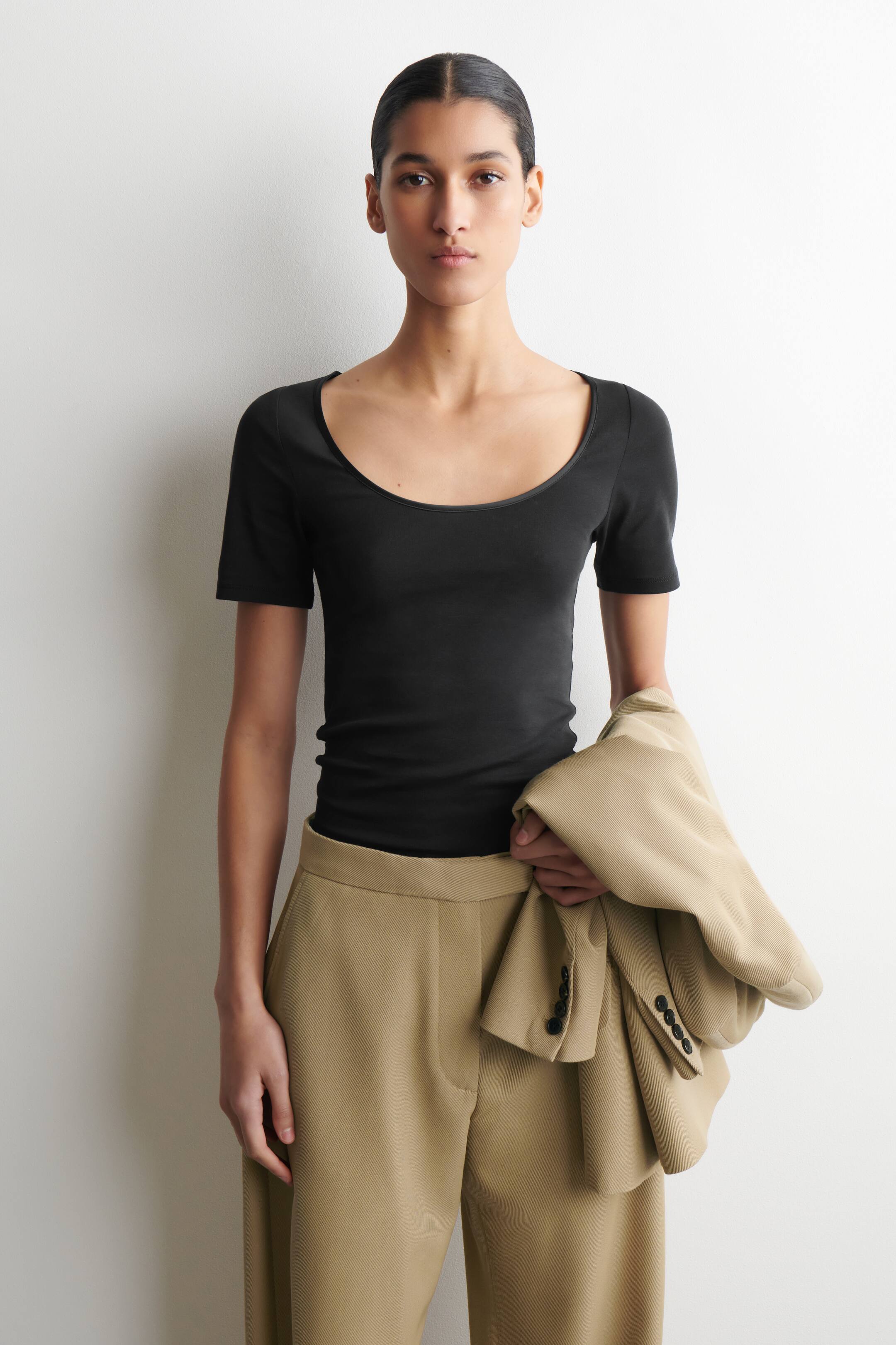 Agrandir l'image: Un haut noir à manches courtes est porté face avant, avec un large col rond et une silhouette ajustée, rentré dans un pantalon beige.