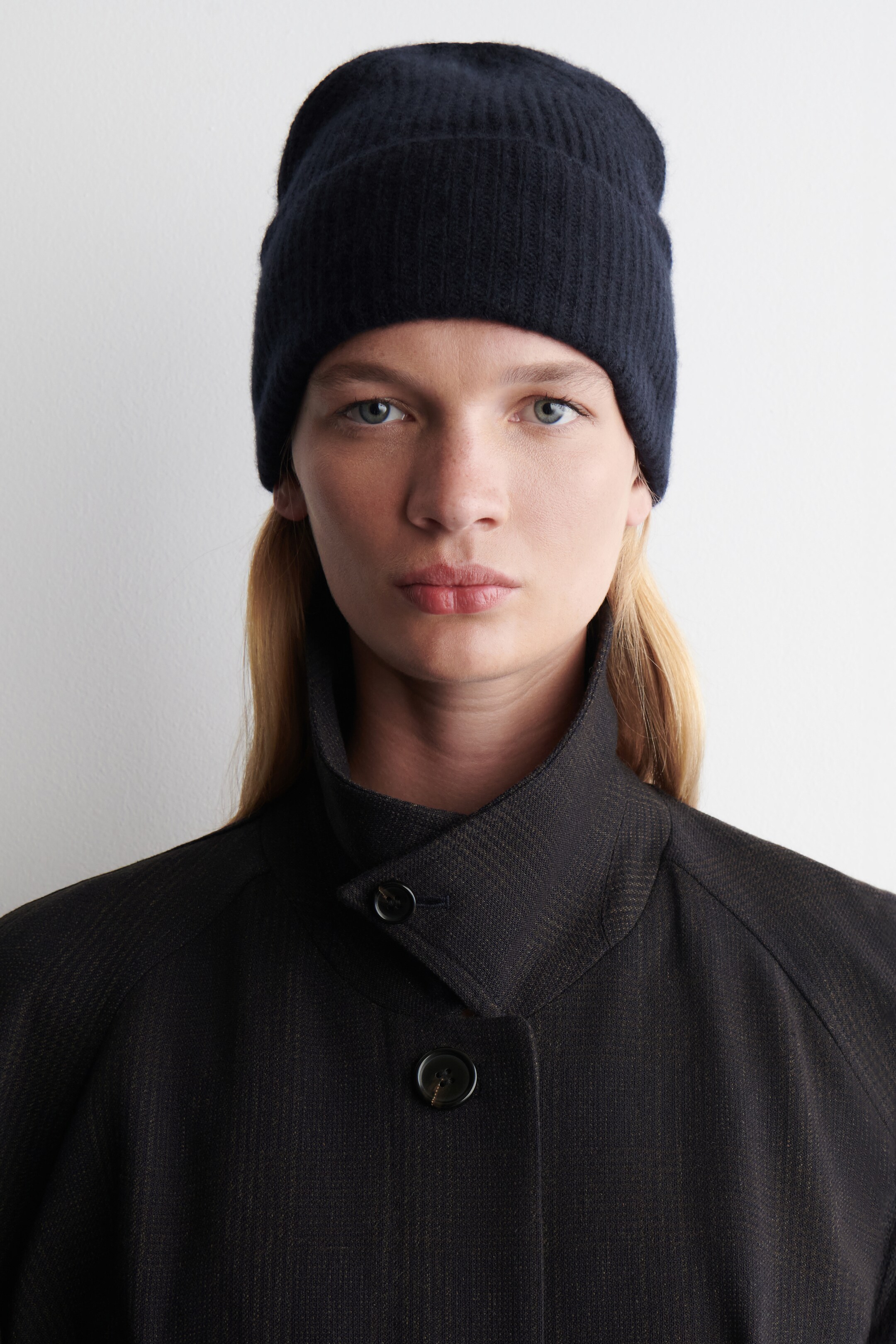 View larger image: A dark navy ribbed knit beanie is covering the top of the head. A dark charcoal jacket, featuring a subtle striped pattern and raglan sleeves, is showcasing a standing collar fastened with a visible button.