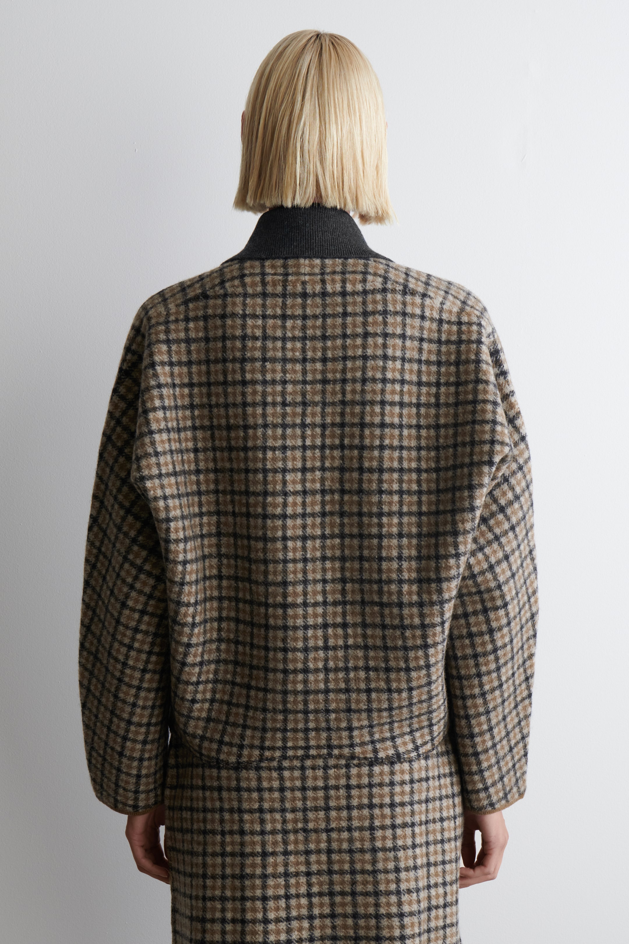 Grösseres Bild ansehen: Eine Jacke und ein passender Rock, beide in einem braun-schwarzen Karomuster, werden von hinten gezeigt. Die Jacke ist mit einer lockeren Passform geschnitten und über einem dunkelgrauen gerippten Rollkragenpullover geschichtet. Der passende Rock reicht unter den Saum der Jacke.