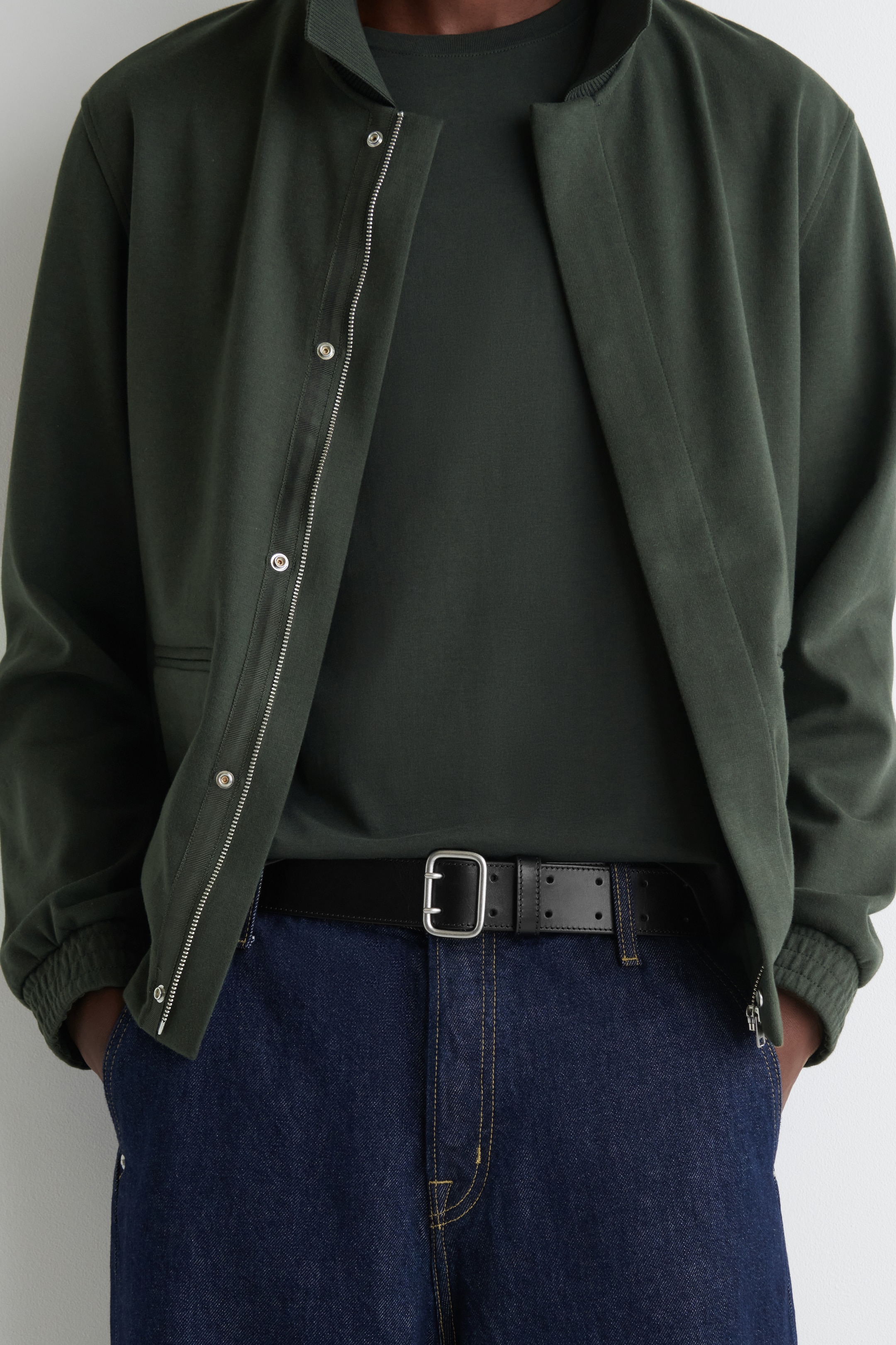 Visualizza immagine più grande: Una giacca bomber verde scuro viene presentata, indossata aperta su una t-shirt verde scuro e jeans blu scuro. La giacca presenta una cerniera argentata e bottoni a pressione sulla sua abbottonatura, un colletto a coste e polsini a coste, mentre una cintura nera con una fibbia a doppia punta argentata stringe i jeans in vita.