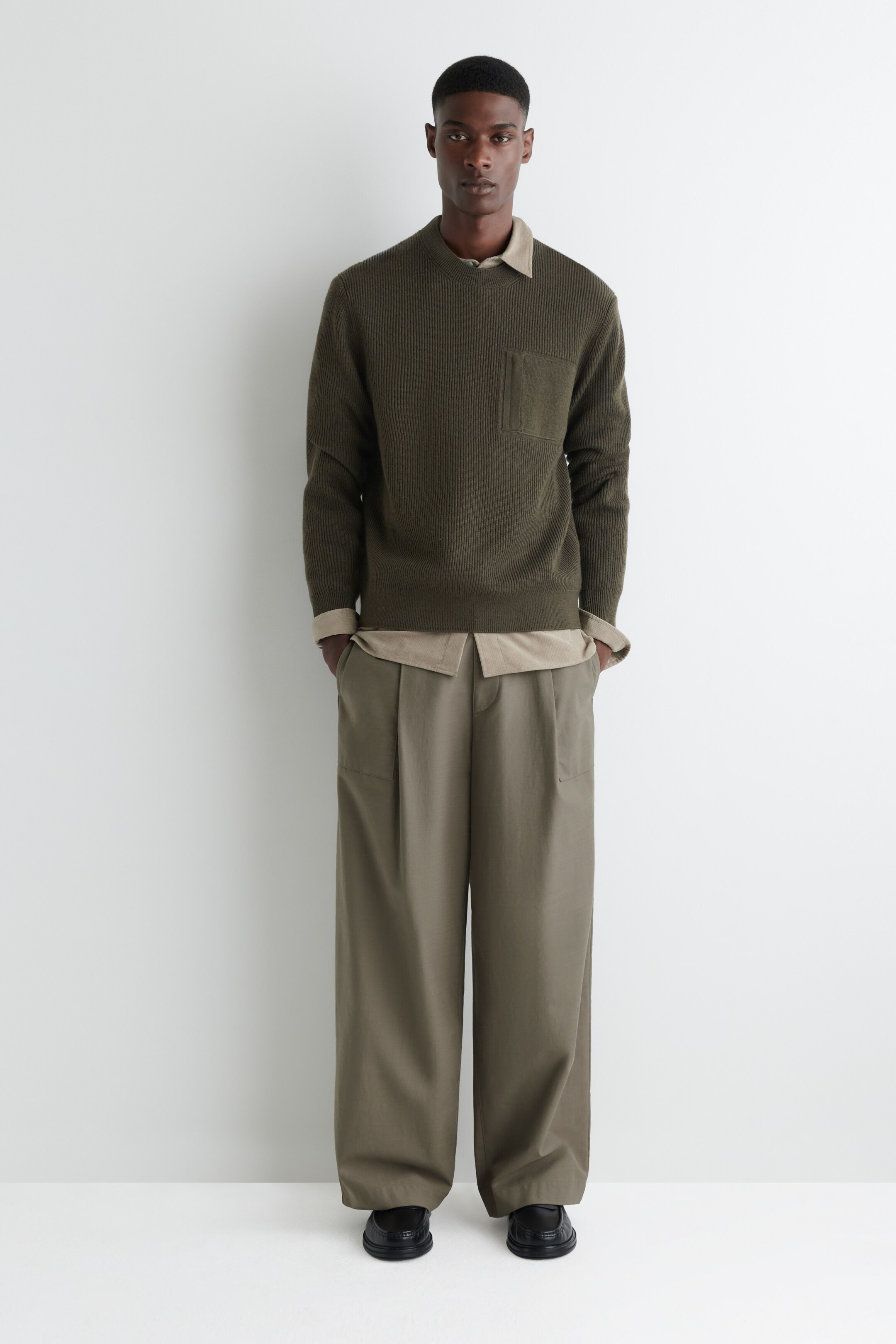 Agrandir l'image: Un pull vert olive est présenté de face, avec un tricot côtelé vertical, un col rond et une poche plaquée sur la poitrine gauche. Il a des poignets et un ourlet côtelés, et est superposé à une chemise à col clair dont le col et les poignets sont visibles.